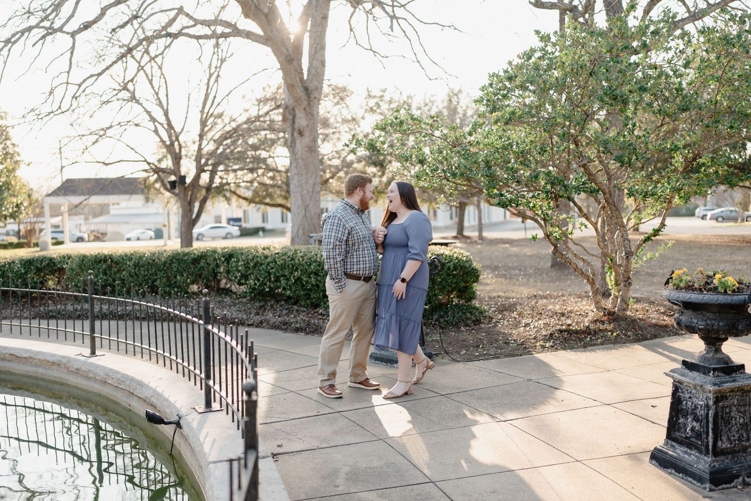 Couple embracing outdoors near a fountain and manicured hedges during a romantic engagement photo session.