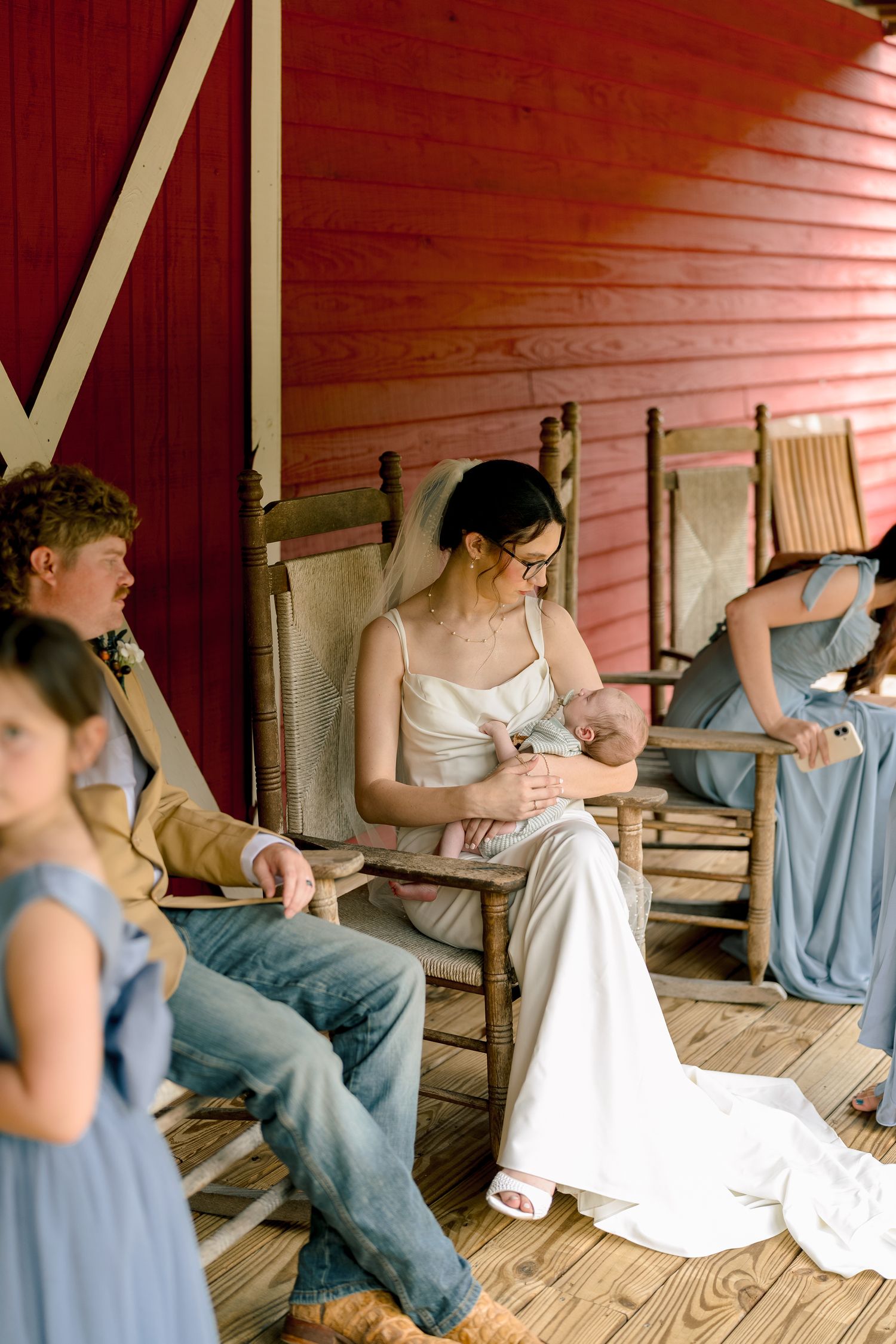 Bride in white gown holding newborn baby while seated in rocking chair on red barn porch.