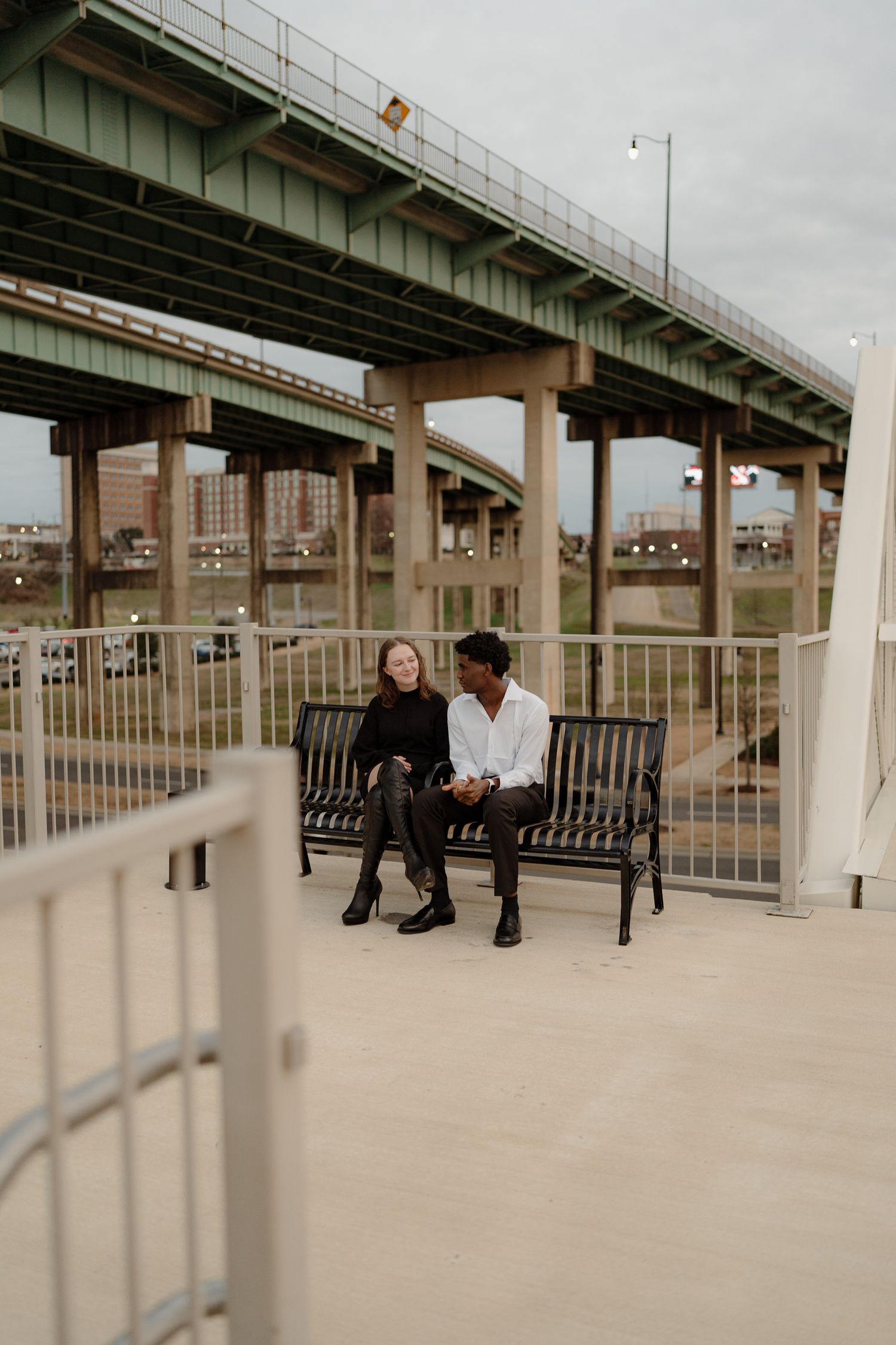 A couple sits together on a black bench beneath a large urban bridge overpass on an overcast day.
