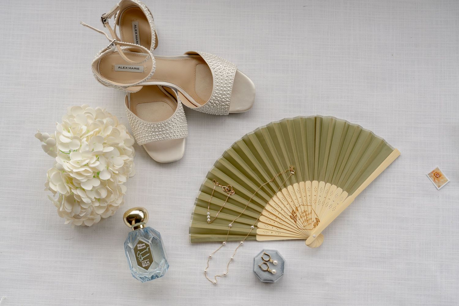 Elegant wedding flat lay with nude heels, green hand fan, white hydrangea, perfume bottle, and rings on white linen.