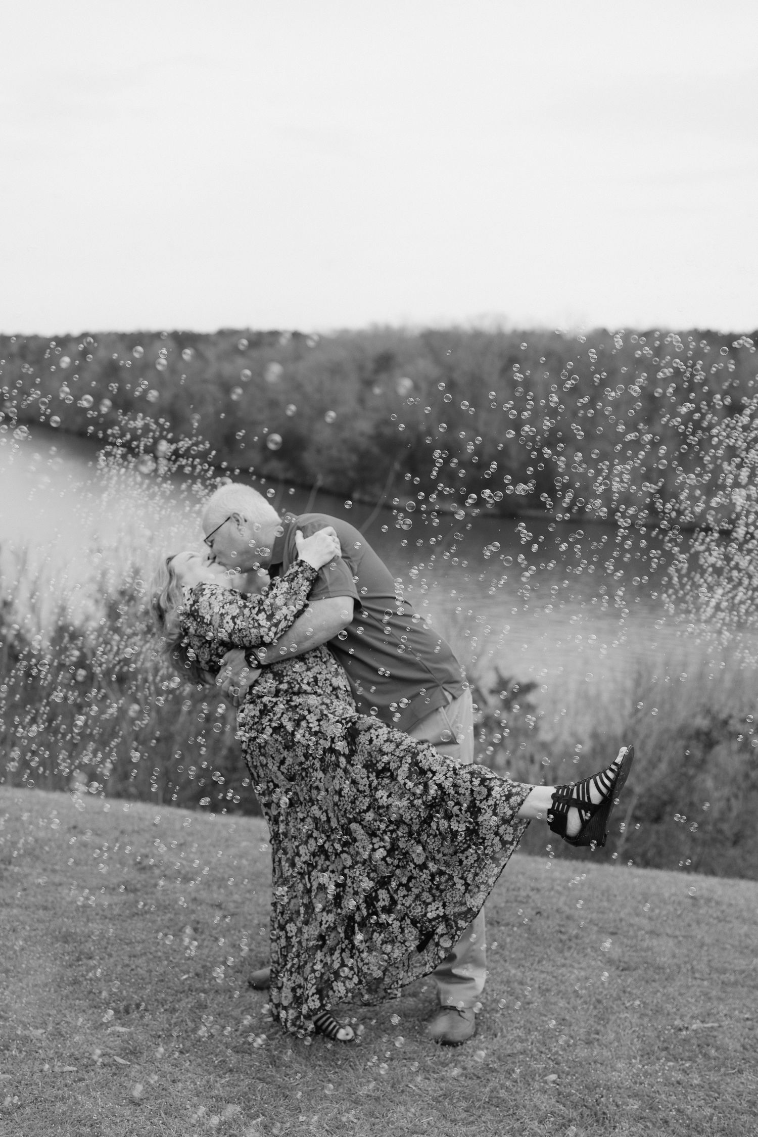 Black and white photo of couple embracing surrounded by bubbles near a scenic waterfront.