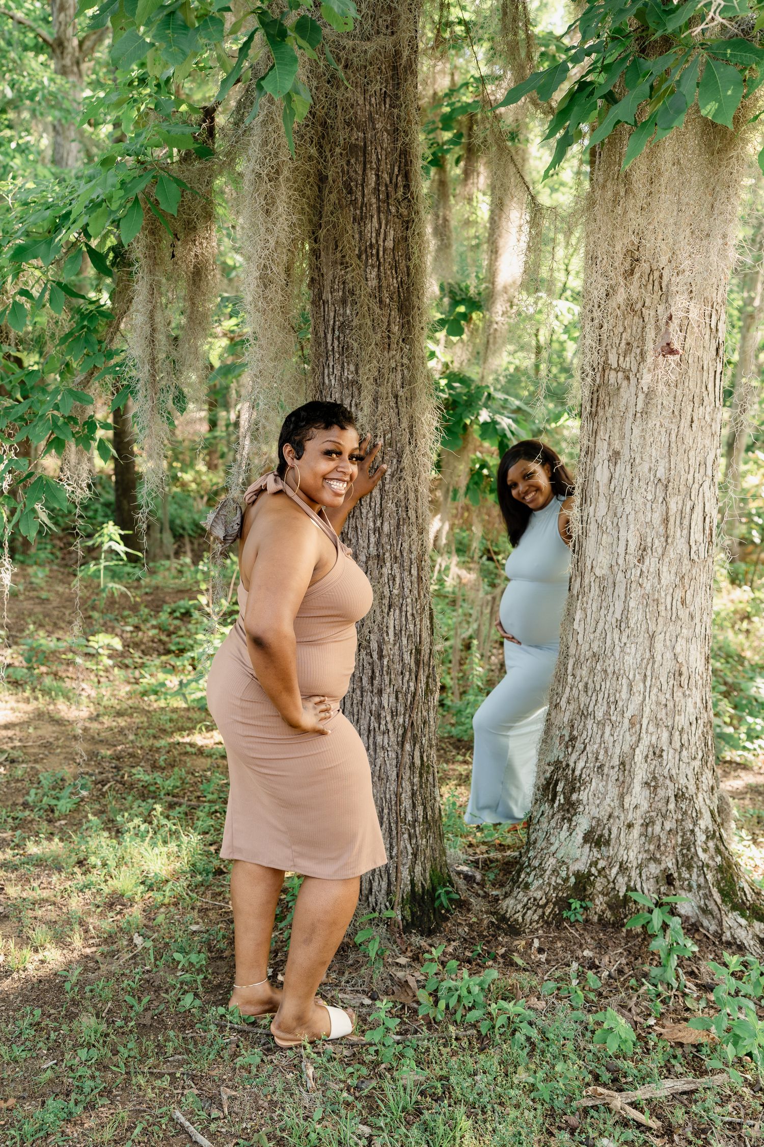 Two women in fitted dresses playfully pose between large tree trunks in a wooded area.