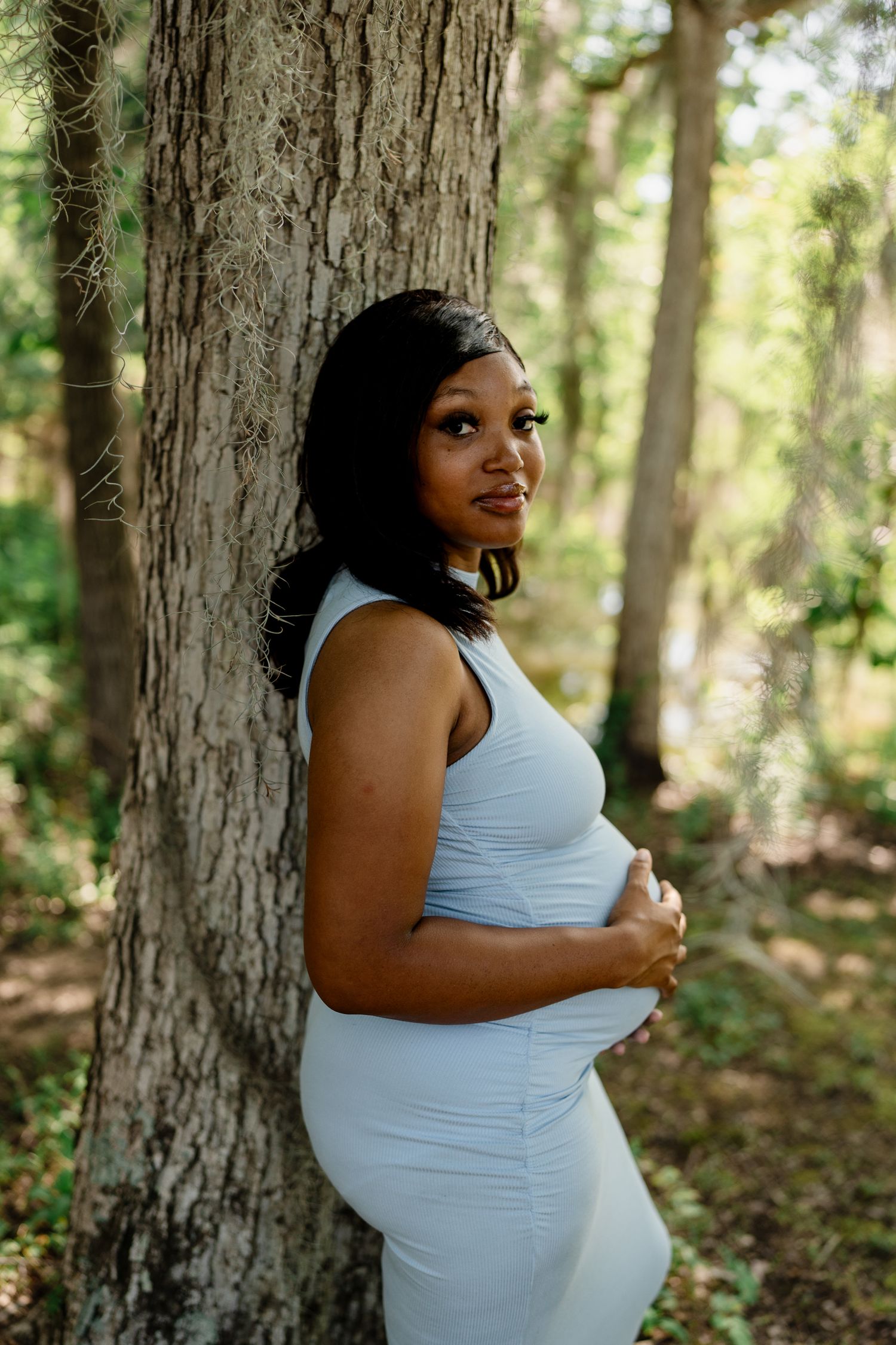 Expectant mother in white dress poses against large tree trunk in sunlit forest.