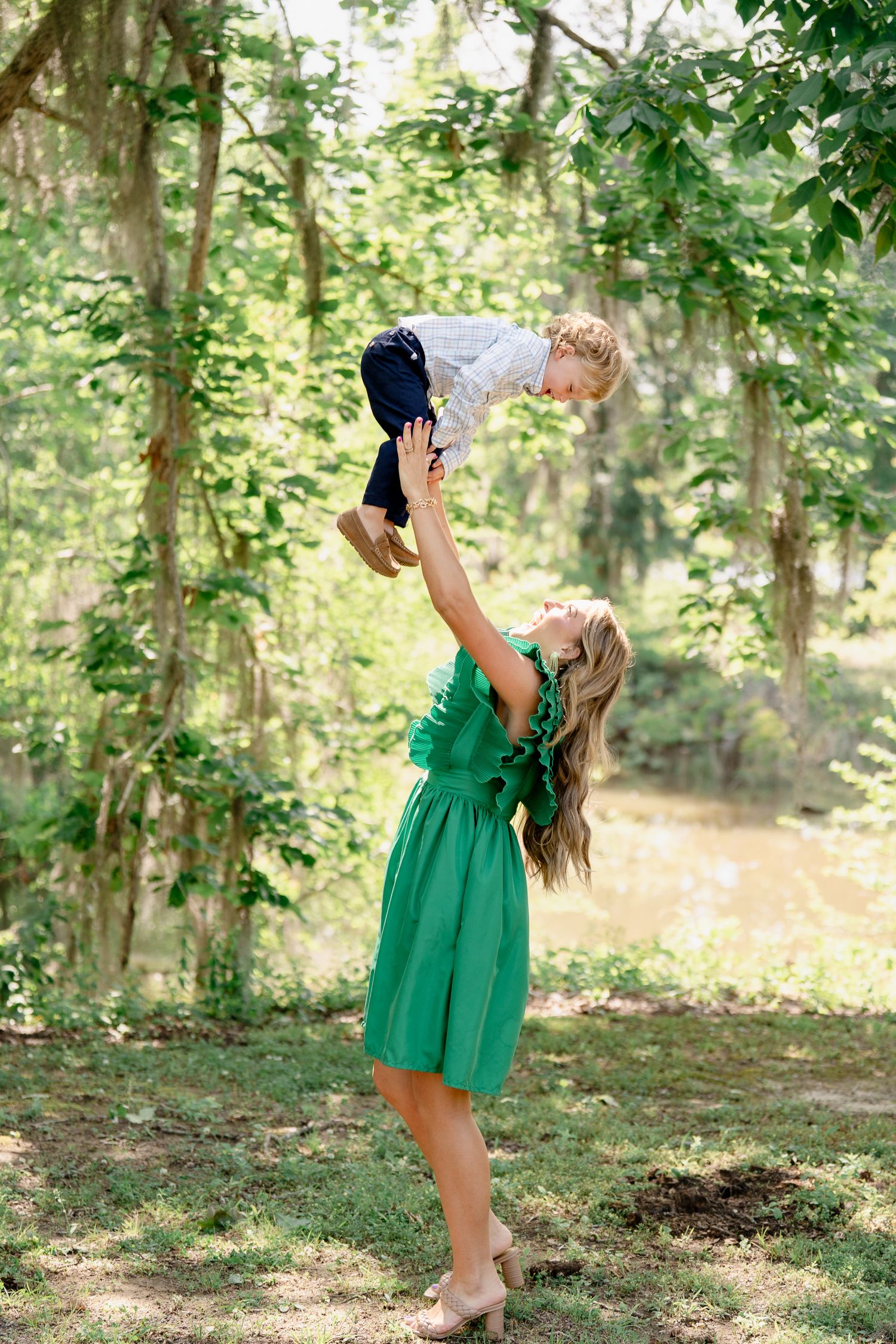 Woman in green dress playing with child in sunlit woodland setting.