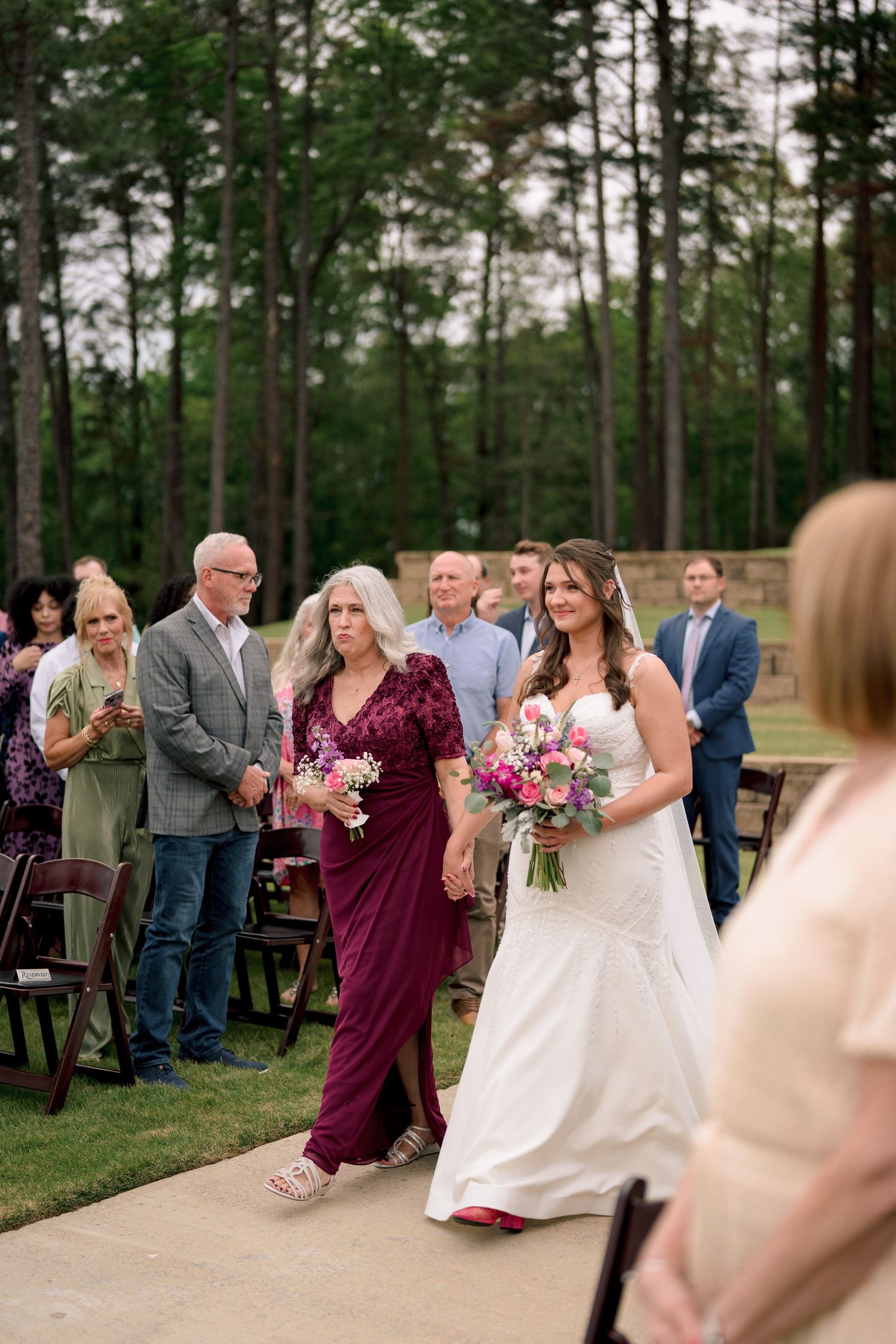 Bride walks down outdoor aisle with mother in burgundy dress, surrounded by standing guests and tall pine trees.