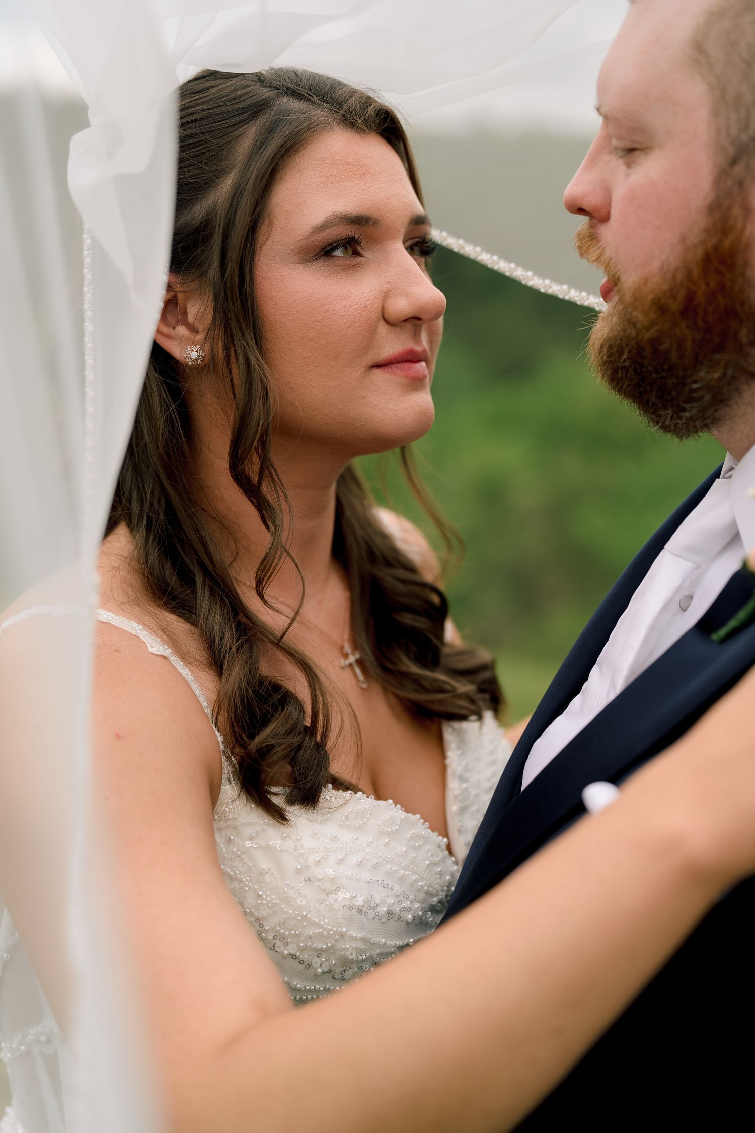 Bride in lace wedding dress gazing lovingly at red-bearded groom under veil during outdoor ceremony.