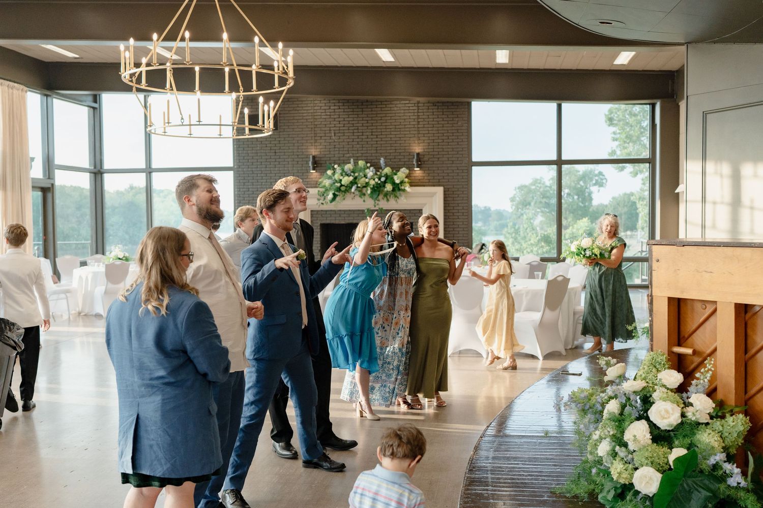 Modern wedding venue interior with chandelier, floral arrangements and large windows overlooking greenery.