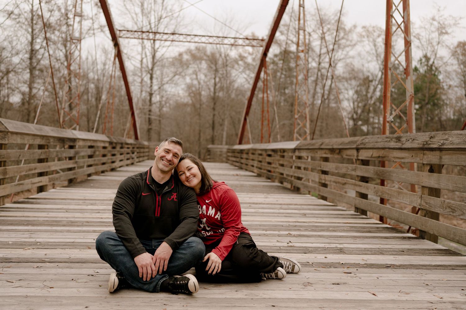 A couple sits close together on a wooden bridge during an outdoor photo session.