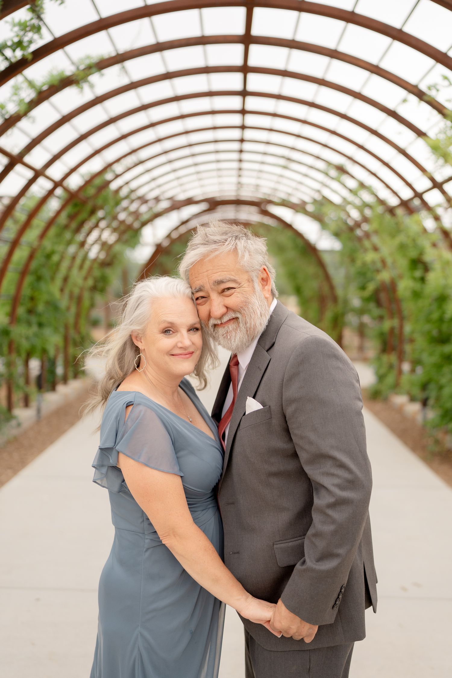 Couple shares intimate moments in a greenhouse walkway with arched wooden beams and natural lighting.