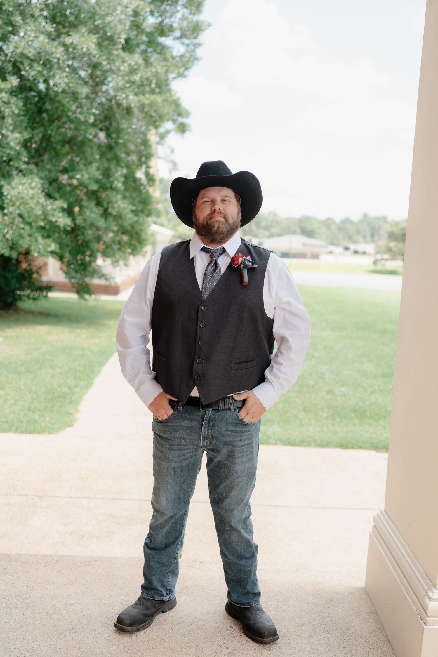 A person in western attire with cowboy hat, vest and jeans stands on a covered porch with trees in background.