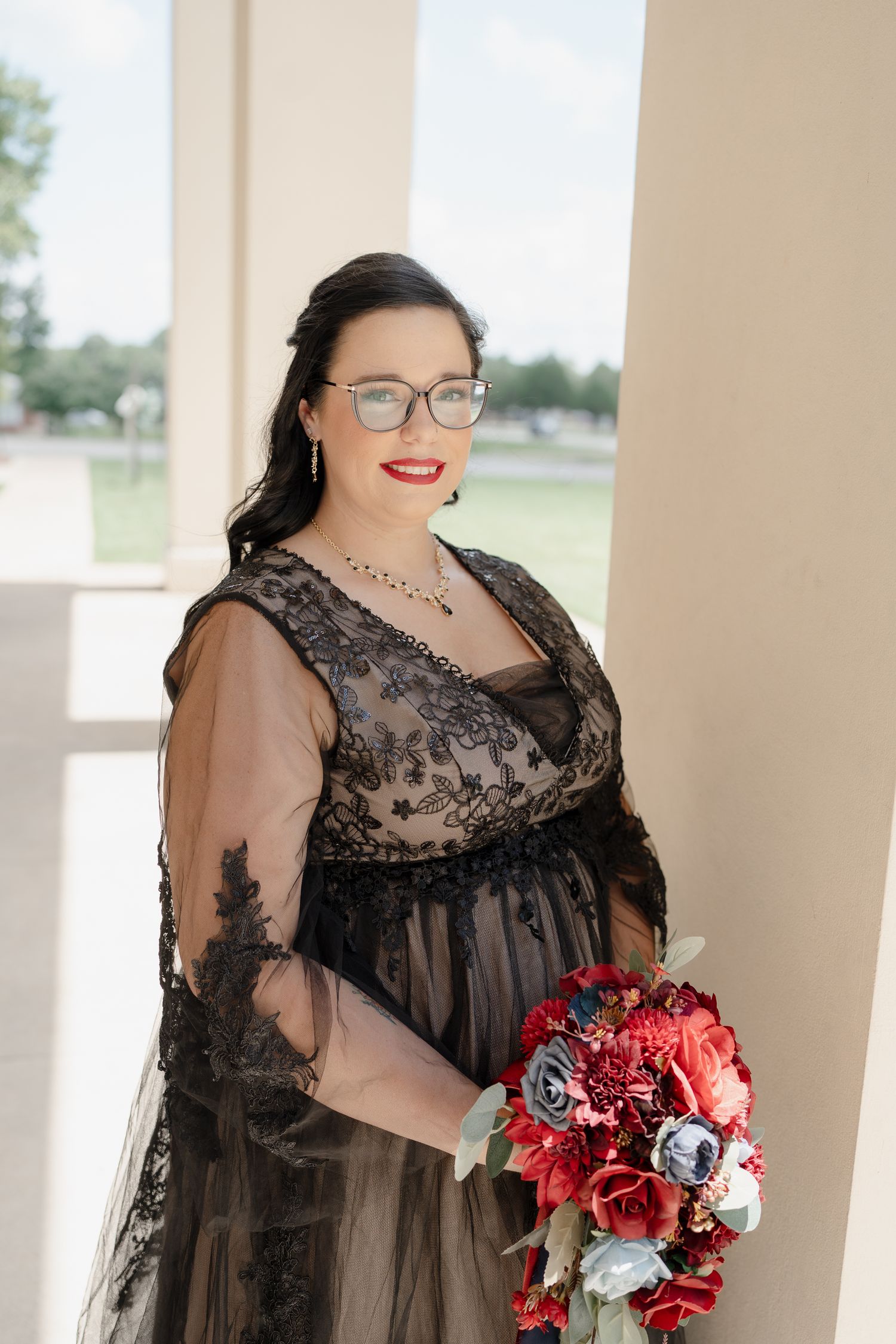 Plus size model in black lace dress poses with red and white floral bouquet in elegant outdoor setting.
