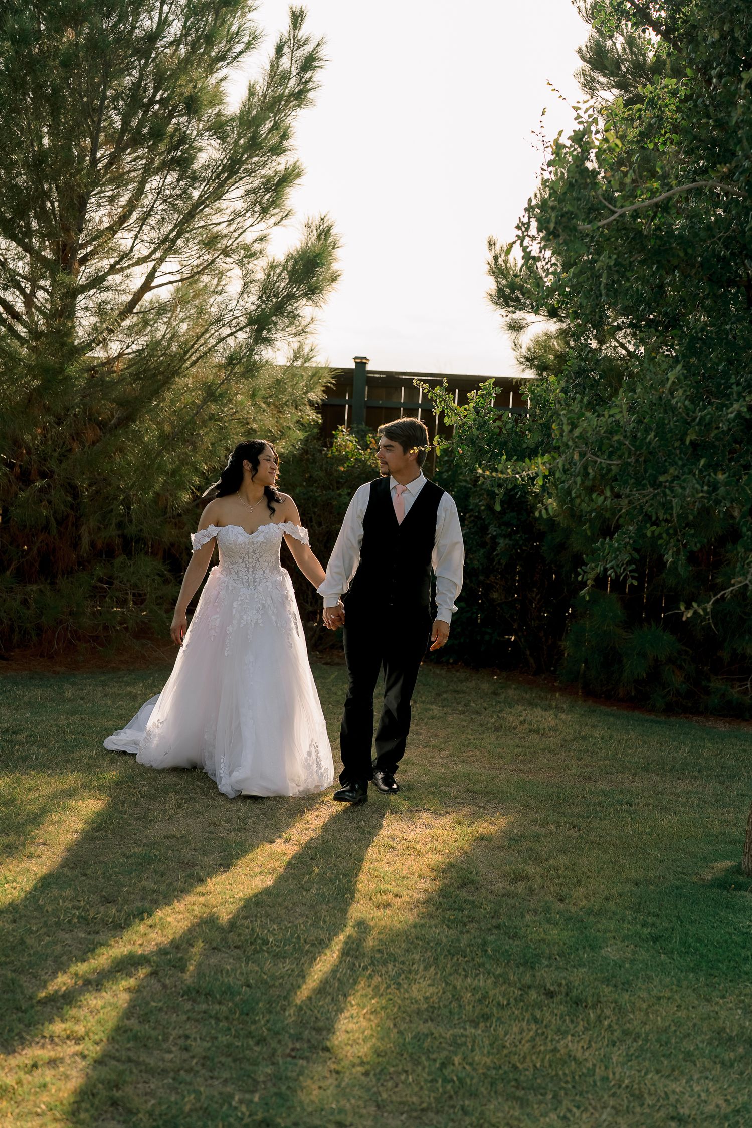 Bride in off-shoulder lace gown and groom in black vest hold hands walking through lush garden at golden hour.