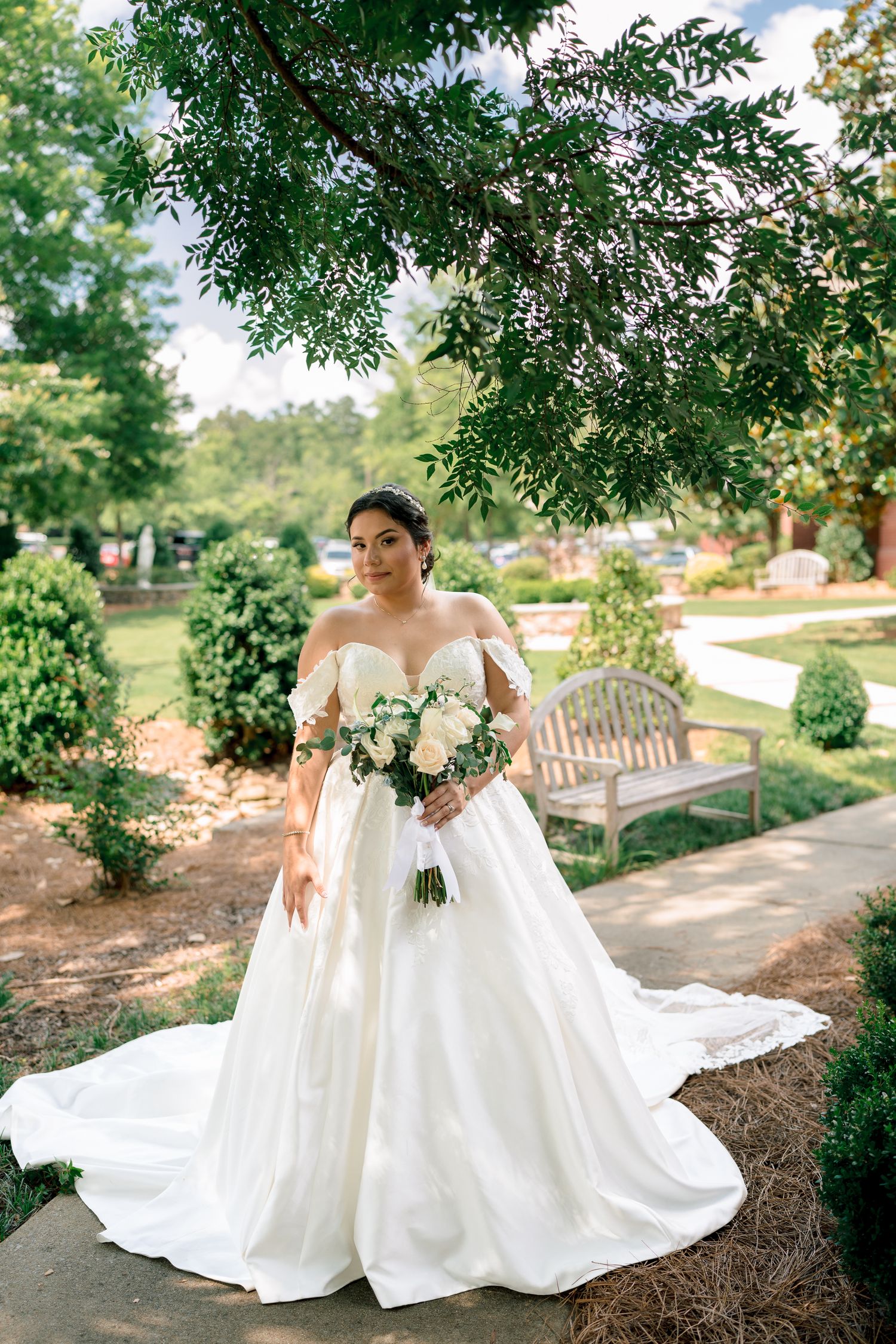 A flowing white wedding dress is elegantly displayed in a garden setting with lush greenery and a stone pathway.