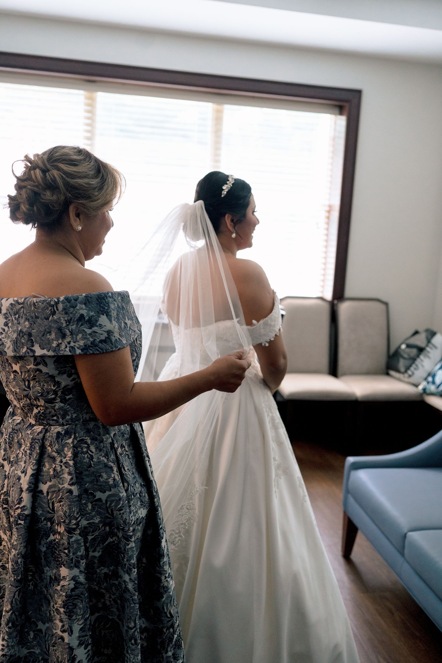 A person in a blue floral dress helps adjust a flowing white wedding gown in a sunlit room.