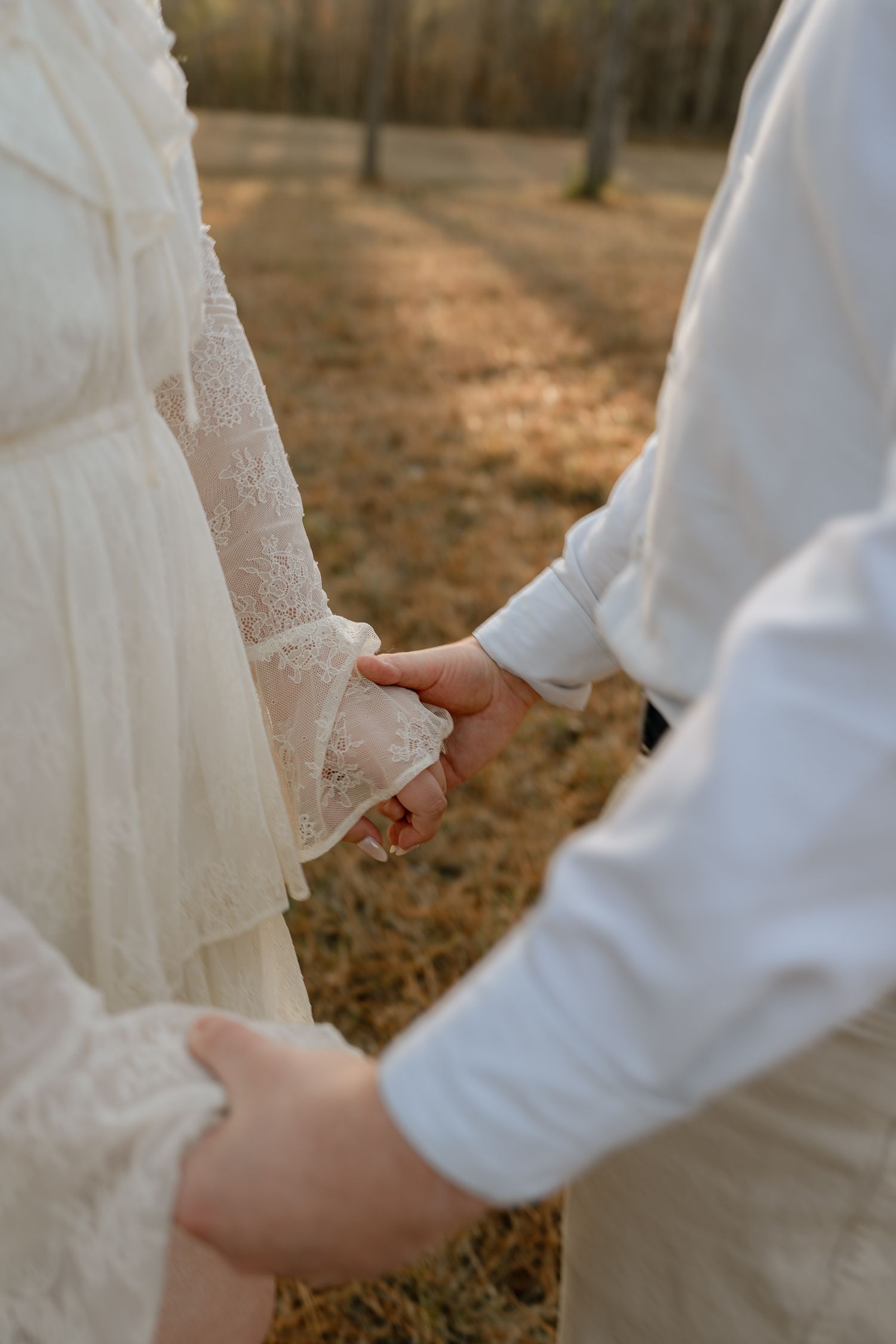 Close-up of couple holding hands in a sunlit autumn forest, wearing white lace and dress shirt.