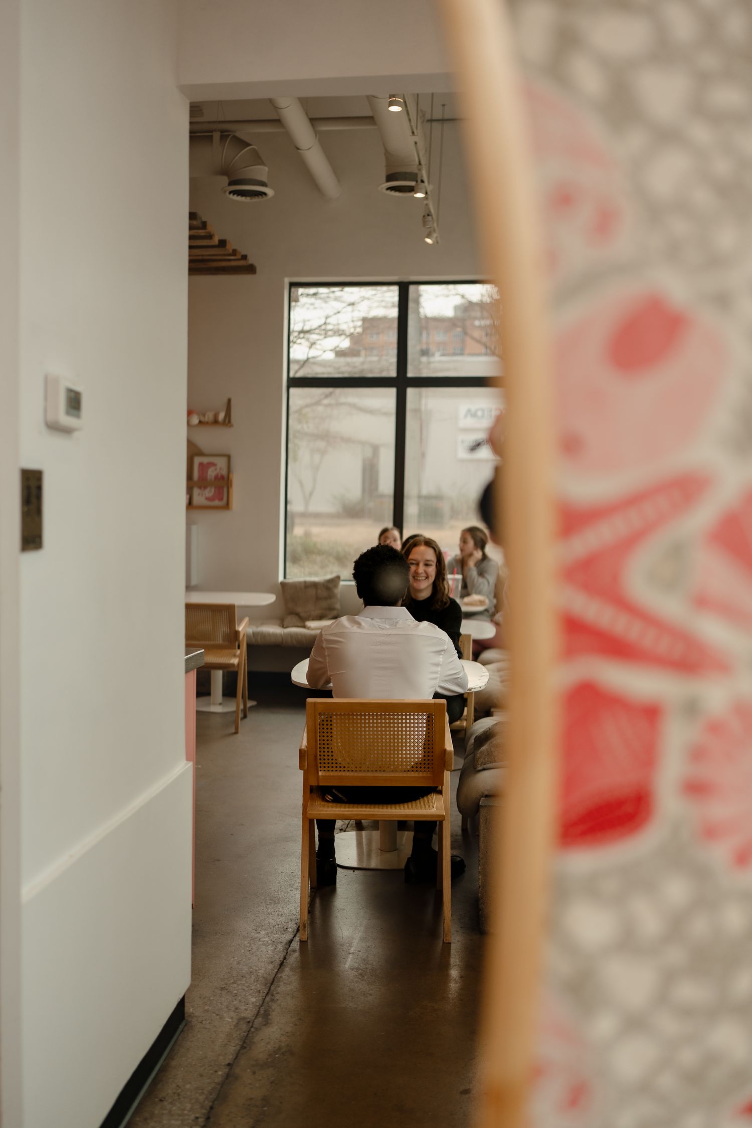 Two people share a warm conversation at a wooden table inside a modern, light-filled cafe with large windows.