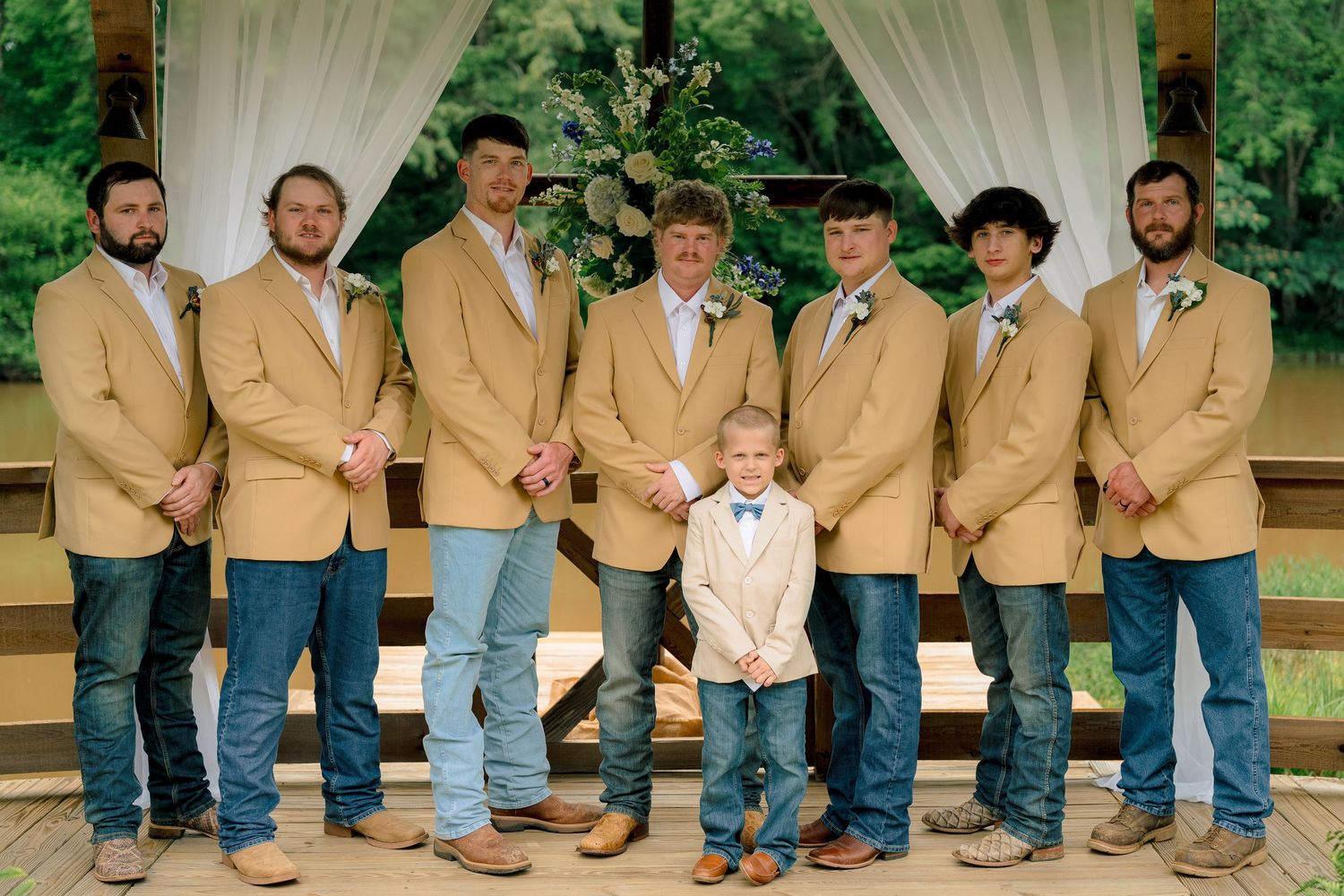 Groom with groomsmen and ring bearer in tan blazers and jeans posing on a wooden outdoor venue deck.