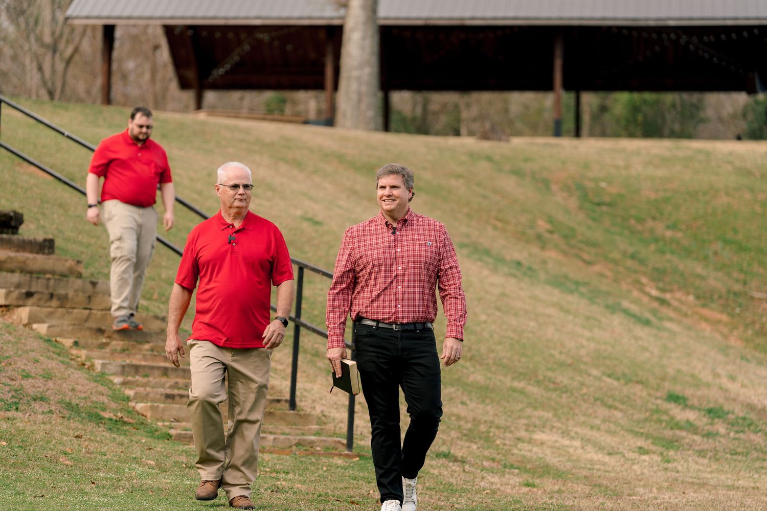 Three men in red polo shirts walking down a grassy hill near a wooden pavilion.