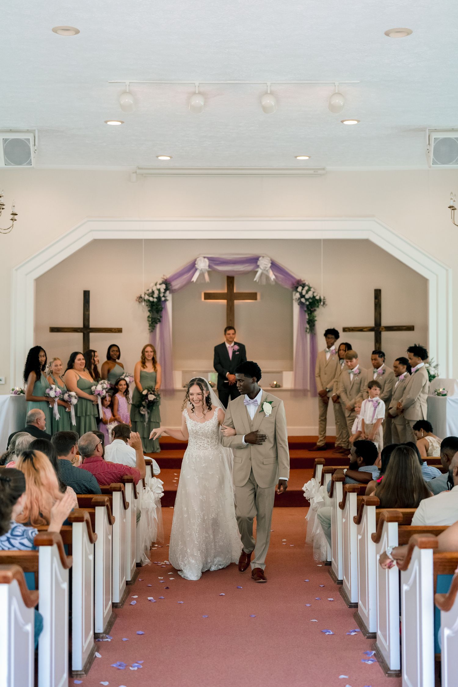 Newlywed interracial couple walks back down church aisle after ceremony, wedding party and guests celebrating in background.