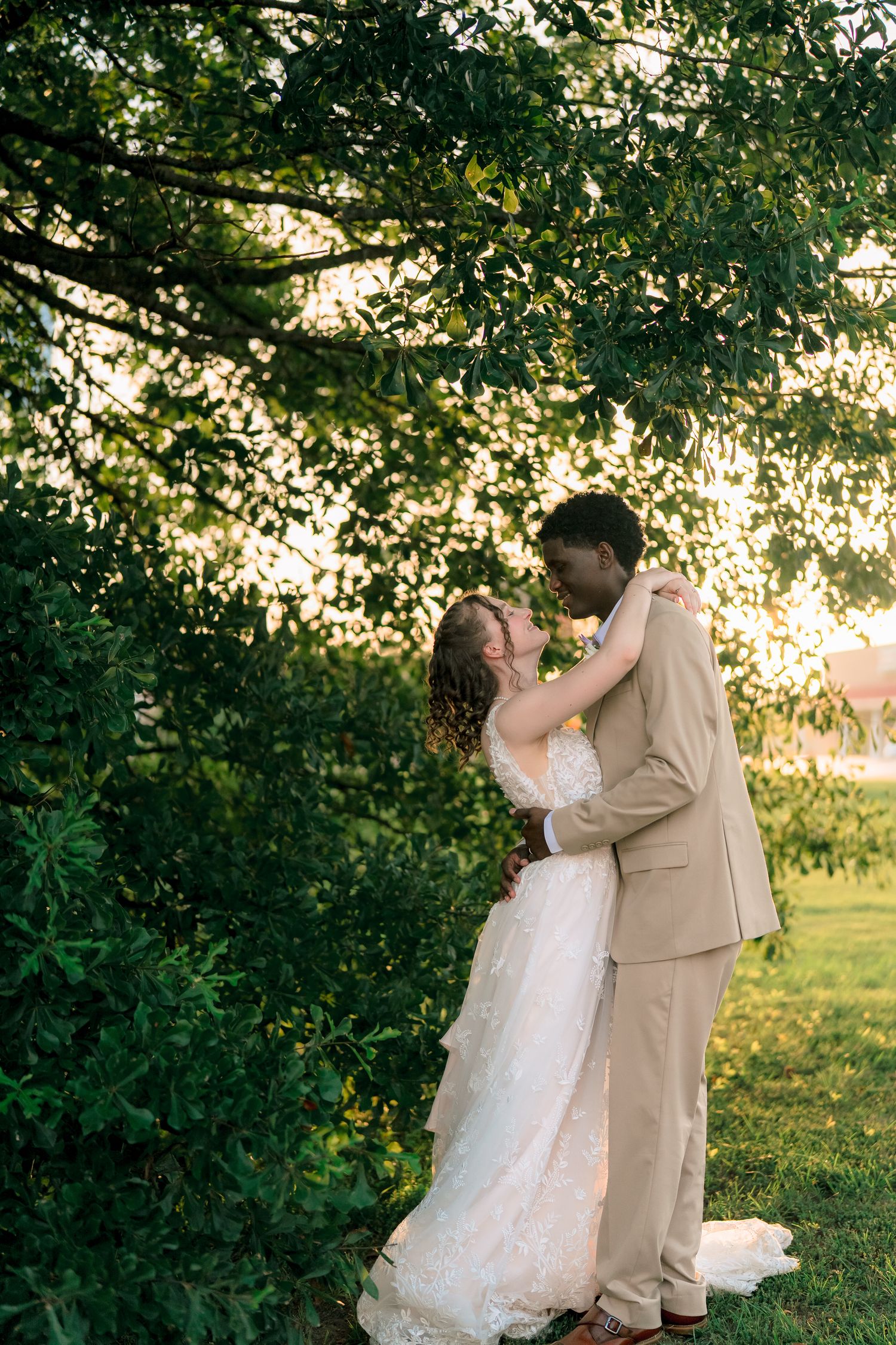 Interracial couple sharing a romantic kiss in wedding attire under lush green trees during golden hour.