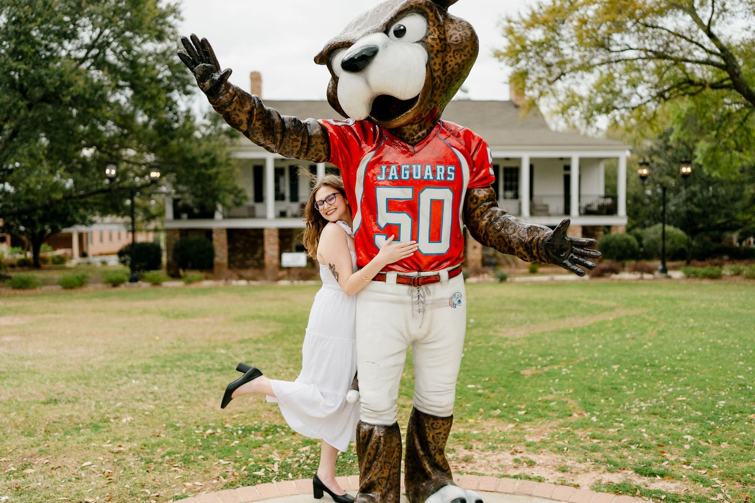 Graduate celebrates with university mascot character wearing sports jersey on campus.