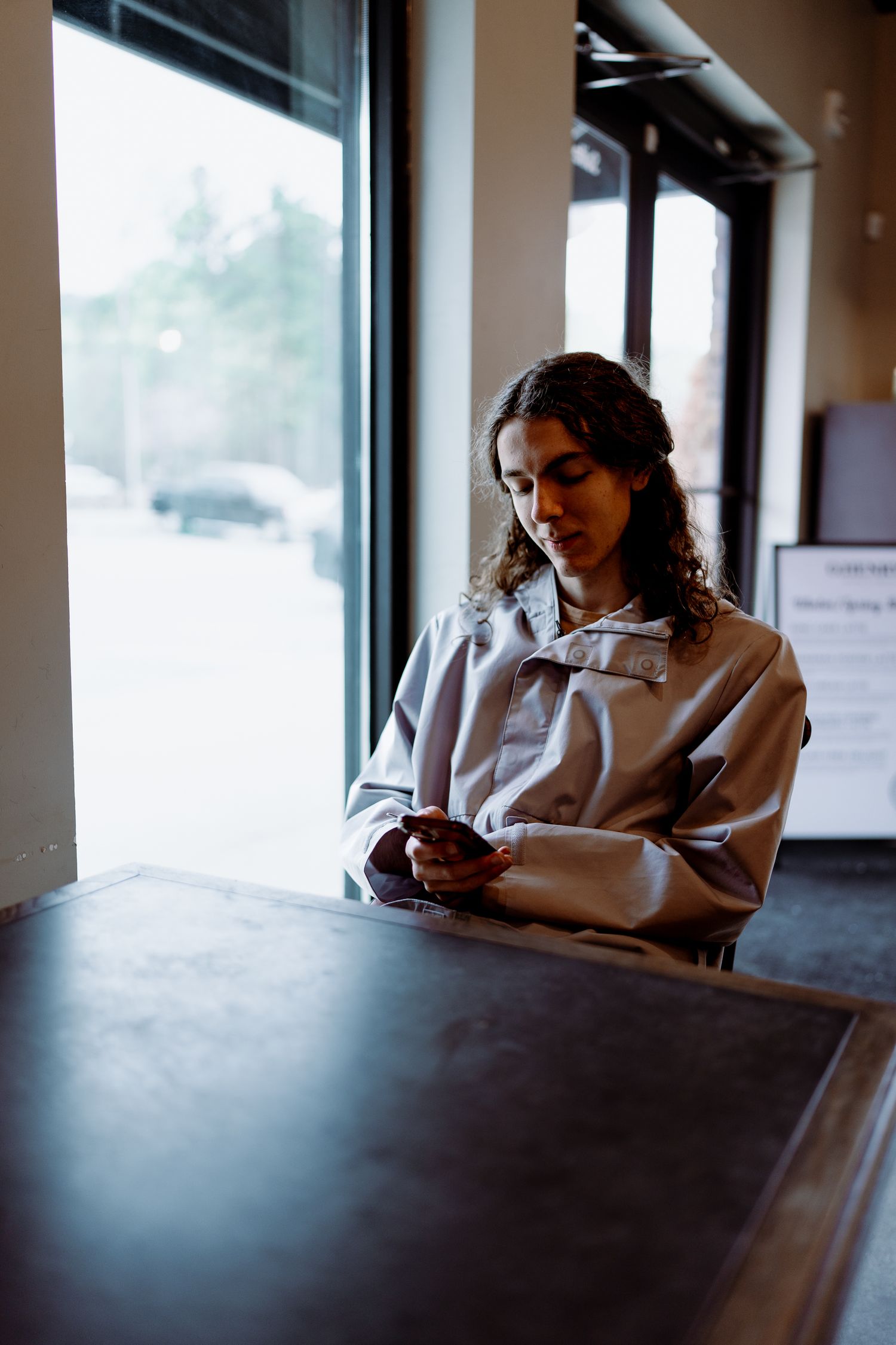 Person in beige jacket checking phone while sitting at dark table by window in moody lighting.