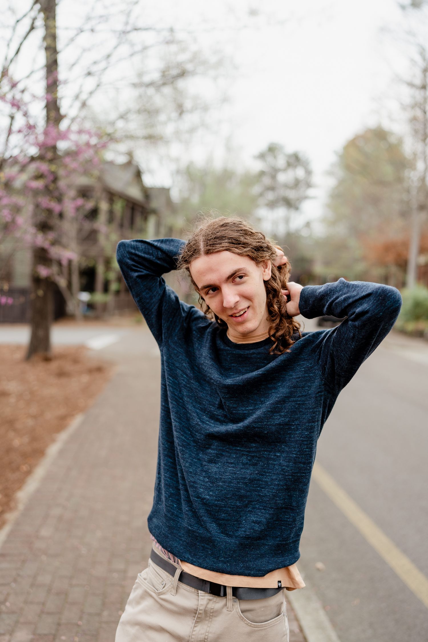 Person in blue sweater poses playfully on brick pathway with blooming trees in background.
