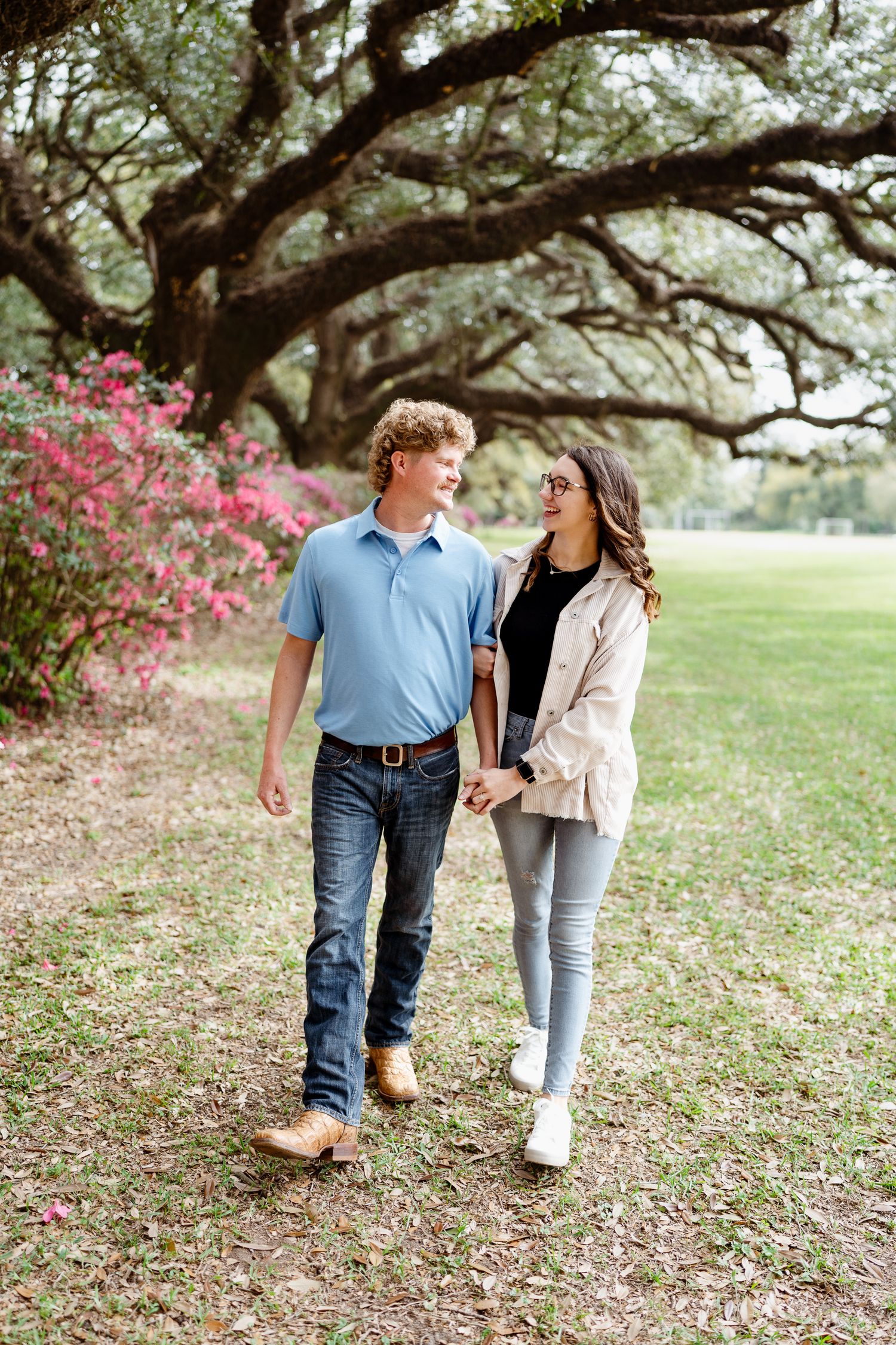 Two people stroll hand in hand beneath sprawling oak trees with pink azaleas in the distance.