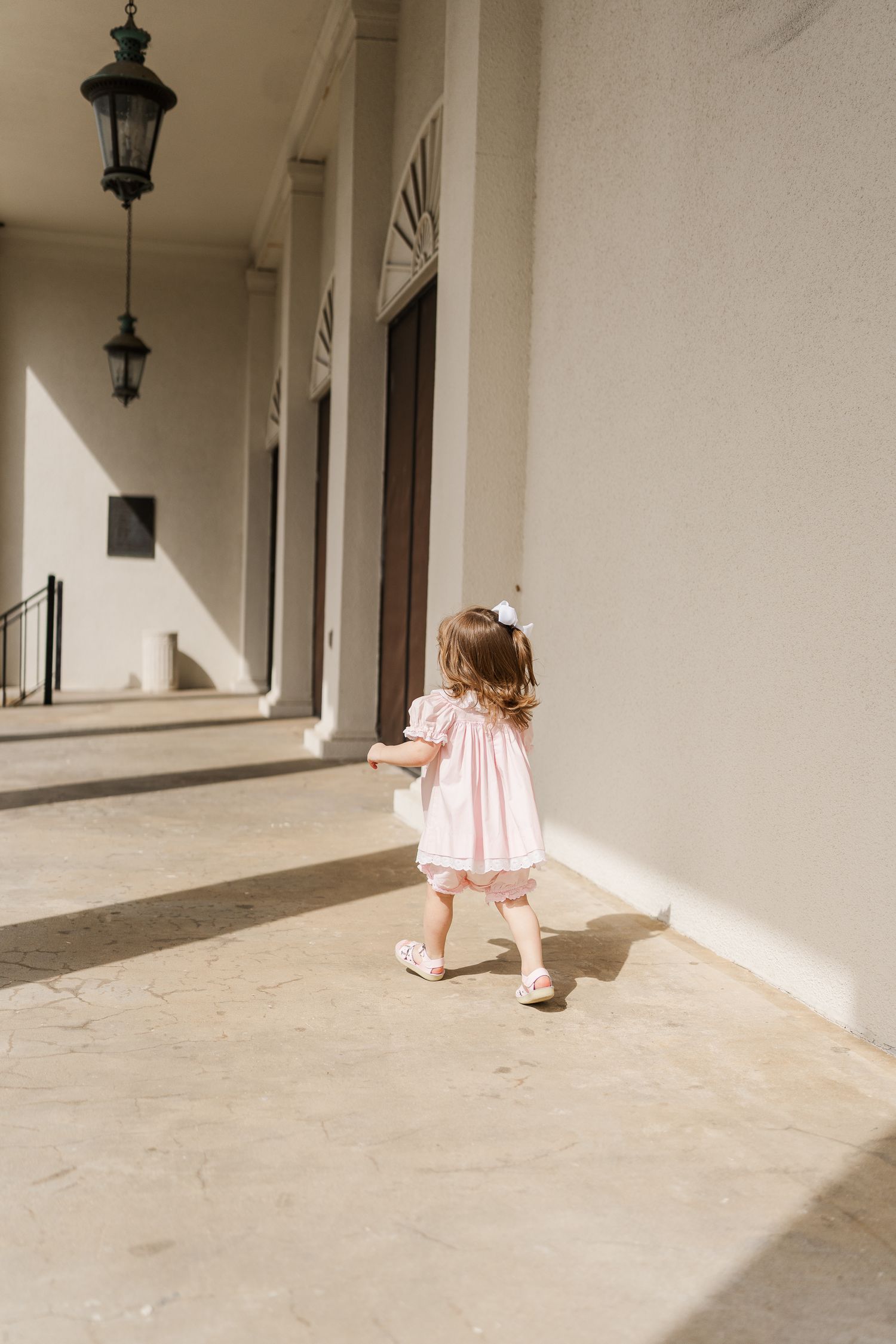A toddler wearing a pink dress walks down a covered walkway with hanging lanterns.