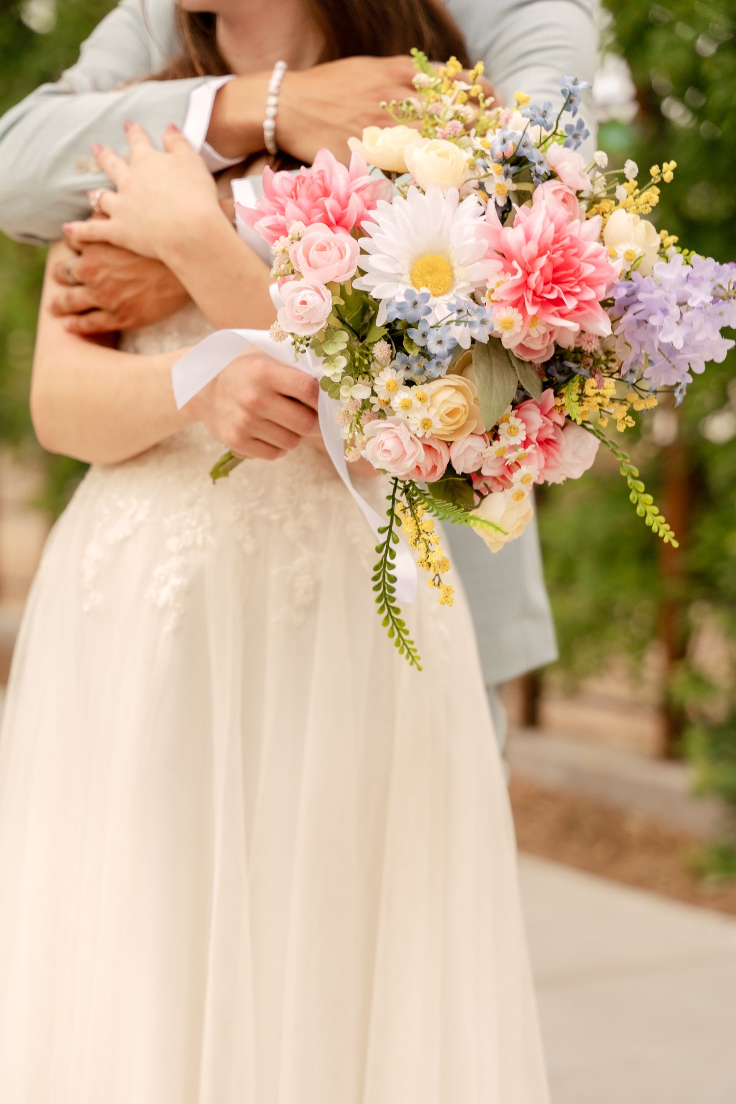 A romantic wedding bouquet with pink dahlias, roses, daisies and purple accents against a flowing white dress.