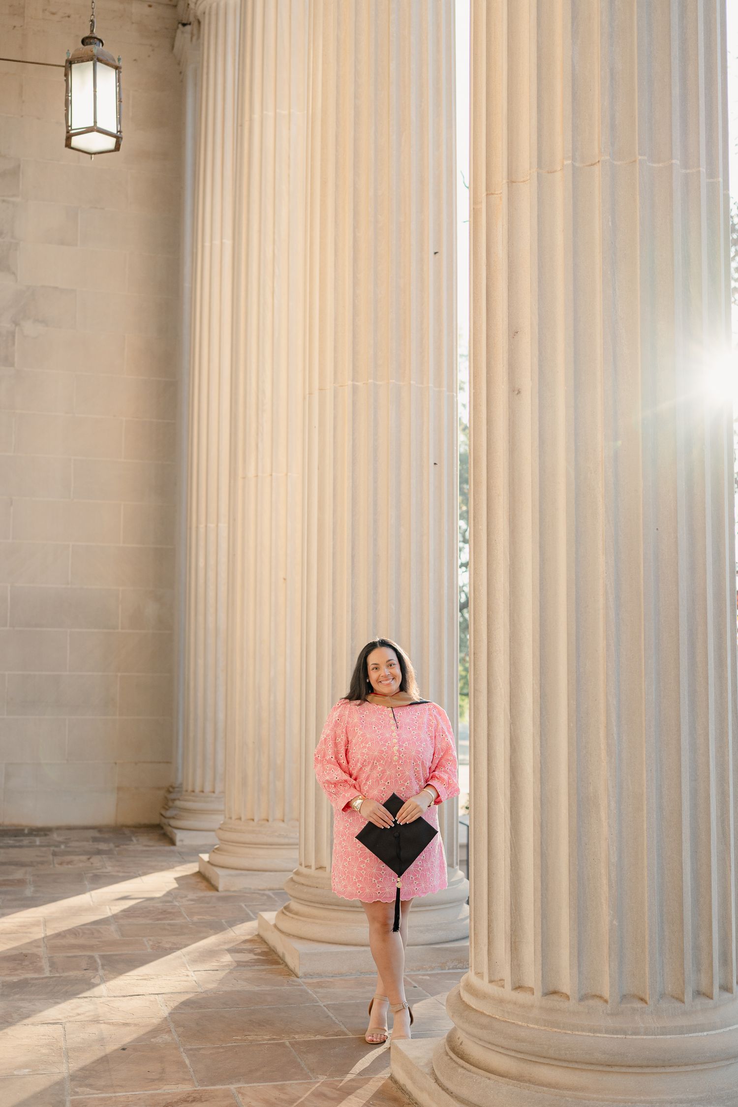A person in a pink dress poses between classical stone columns in an architectural setting with natural lighting.