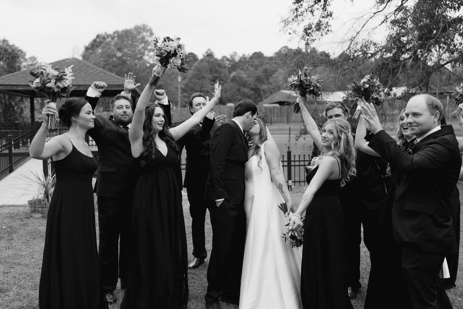 Wedding party poses playfully in a line outdoors with black attire and autumn foliage in the background.