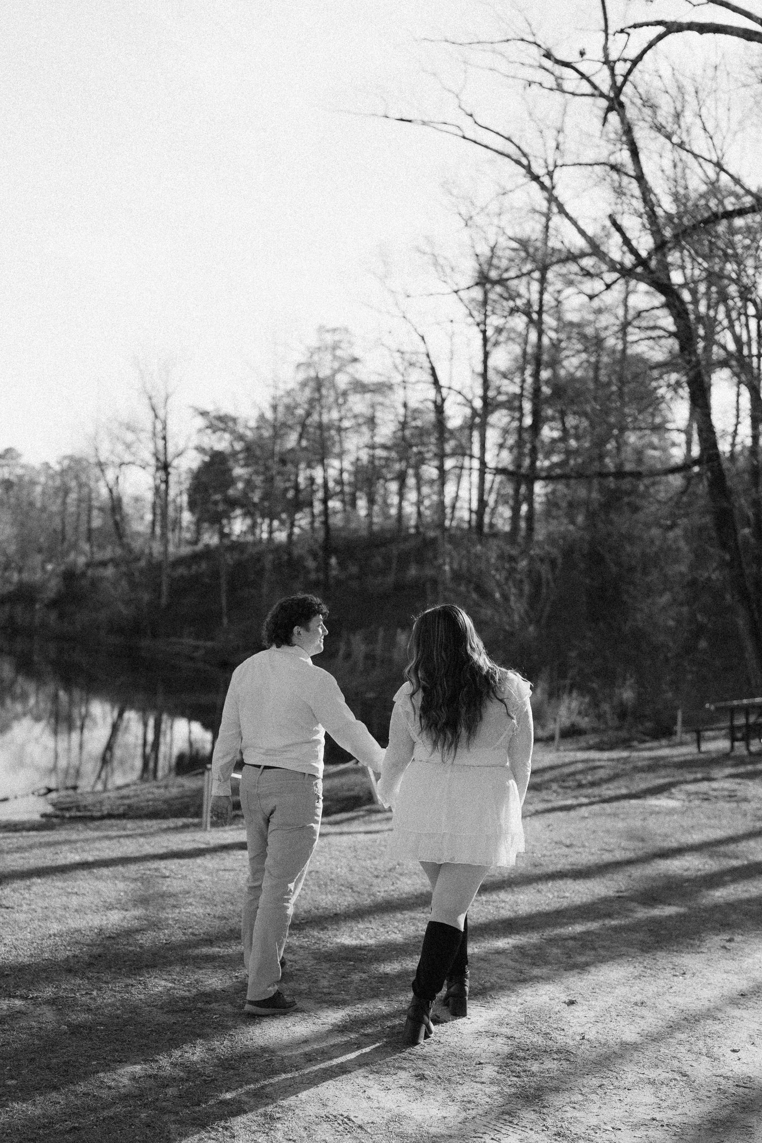 Black and white photo of couple walking hand in hand toward a peaceful lake among tall trees.