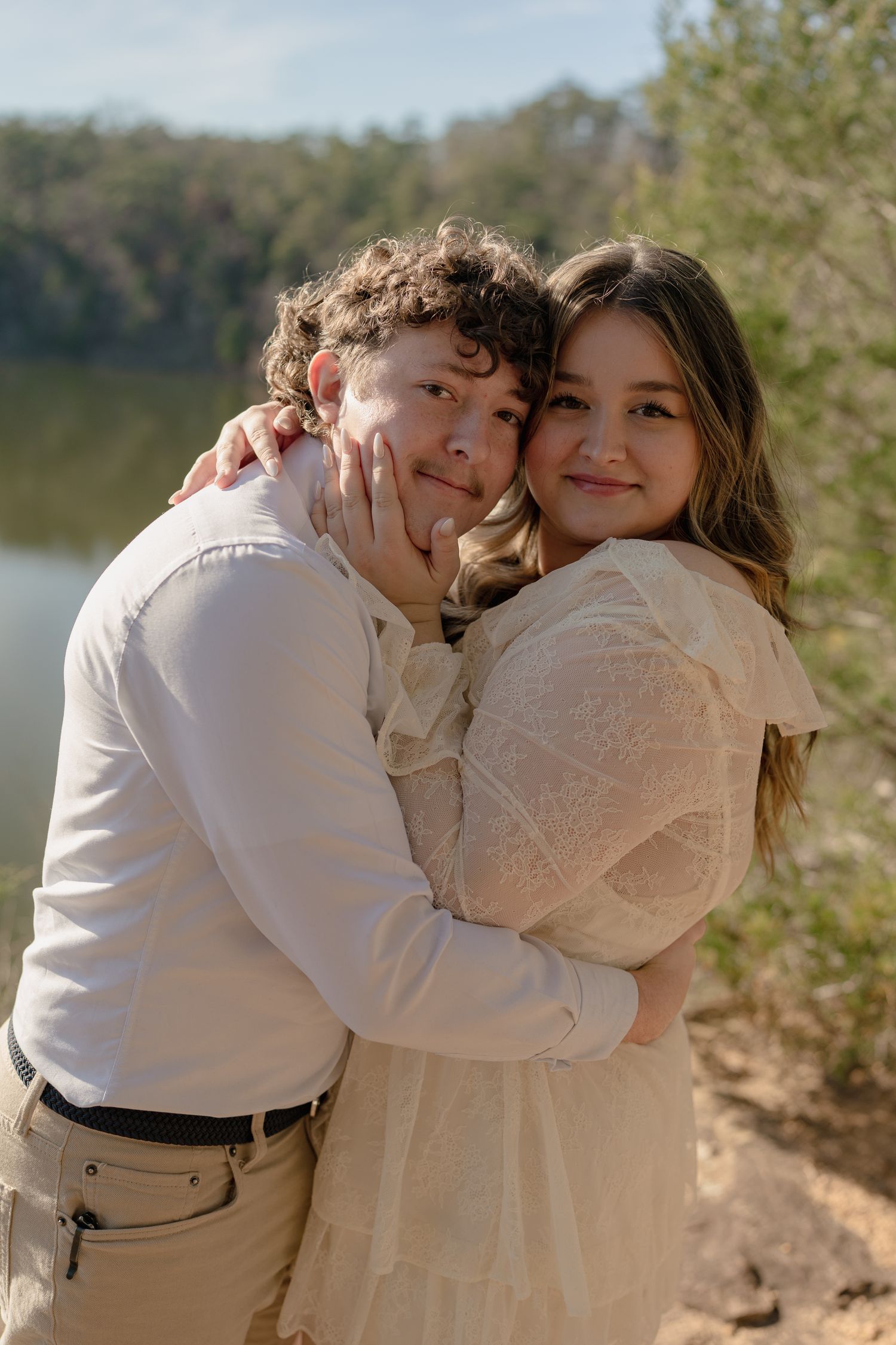 Young couple embracing outdoors near a lake, wearing neutral tones, smiling warmly in a romantic portrait.