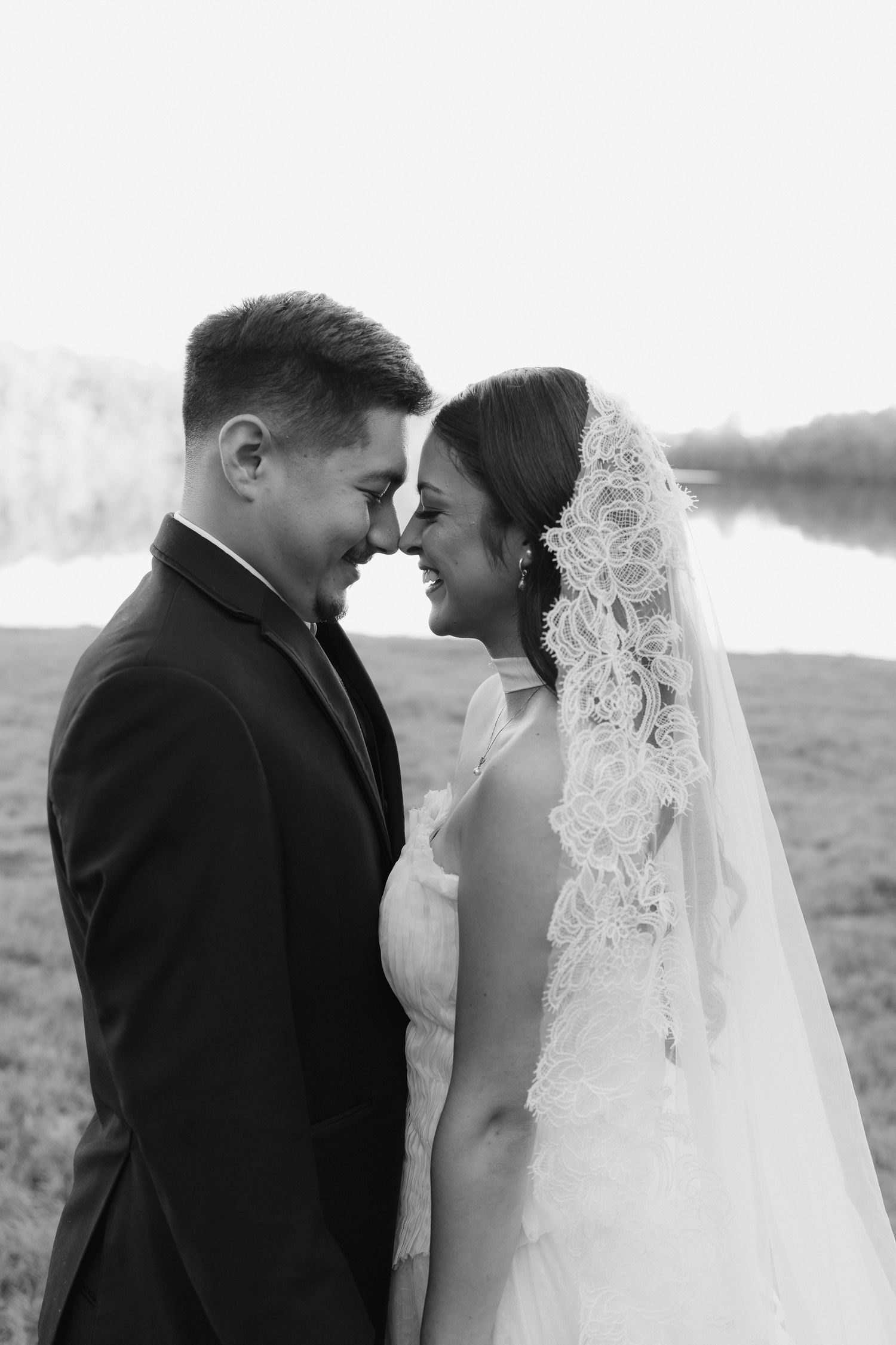Black and white wedding photo of smiling bride in lace veil and groom in suit touching foreheads near a lake.