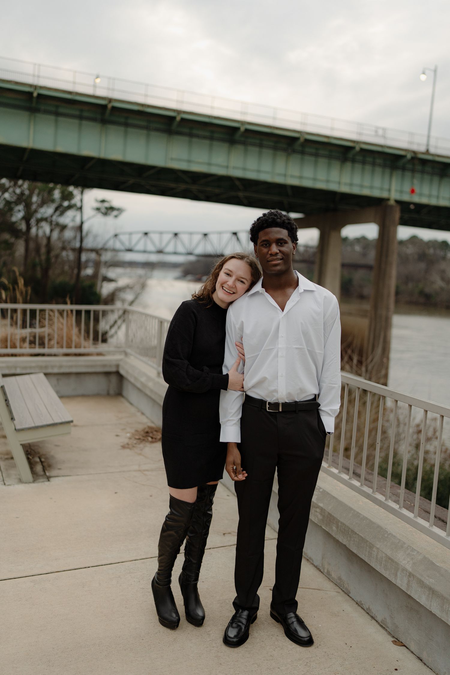 Interracial couple embracing and smiling together on a riverside walkway with a bridge in the background.