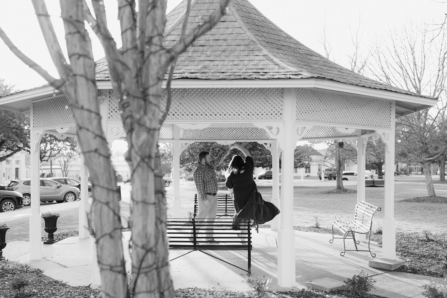 Black and white photo of a couple dancing together inside a white gazebo in a park setting.