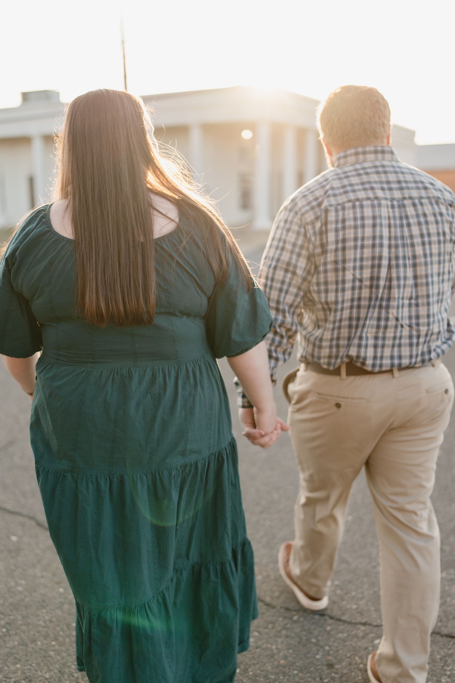 Couple holding hands walking toward a sunset, woman in green dress and man in plaid shirt, viewed from behind.