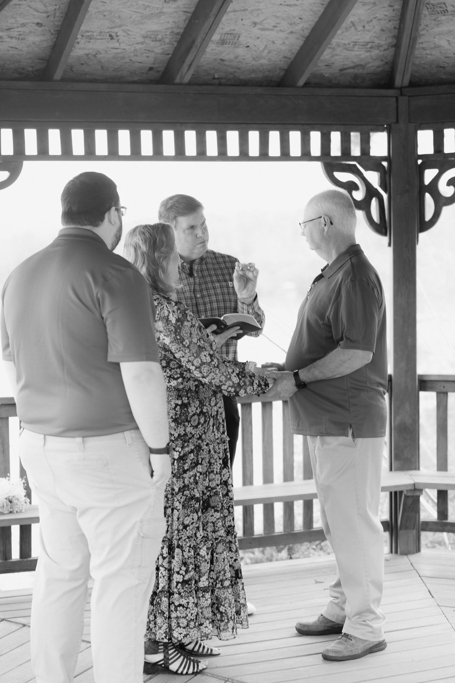 Couple exchanges vows in a wooden gazebo with witnesses in a black and white wedding photo.