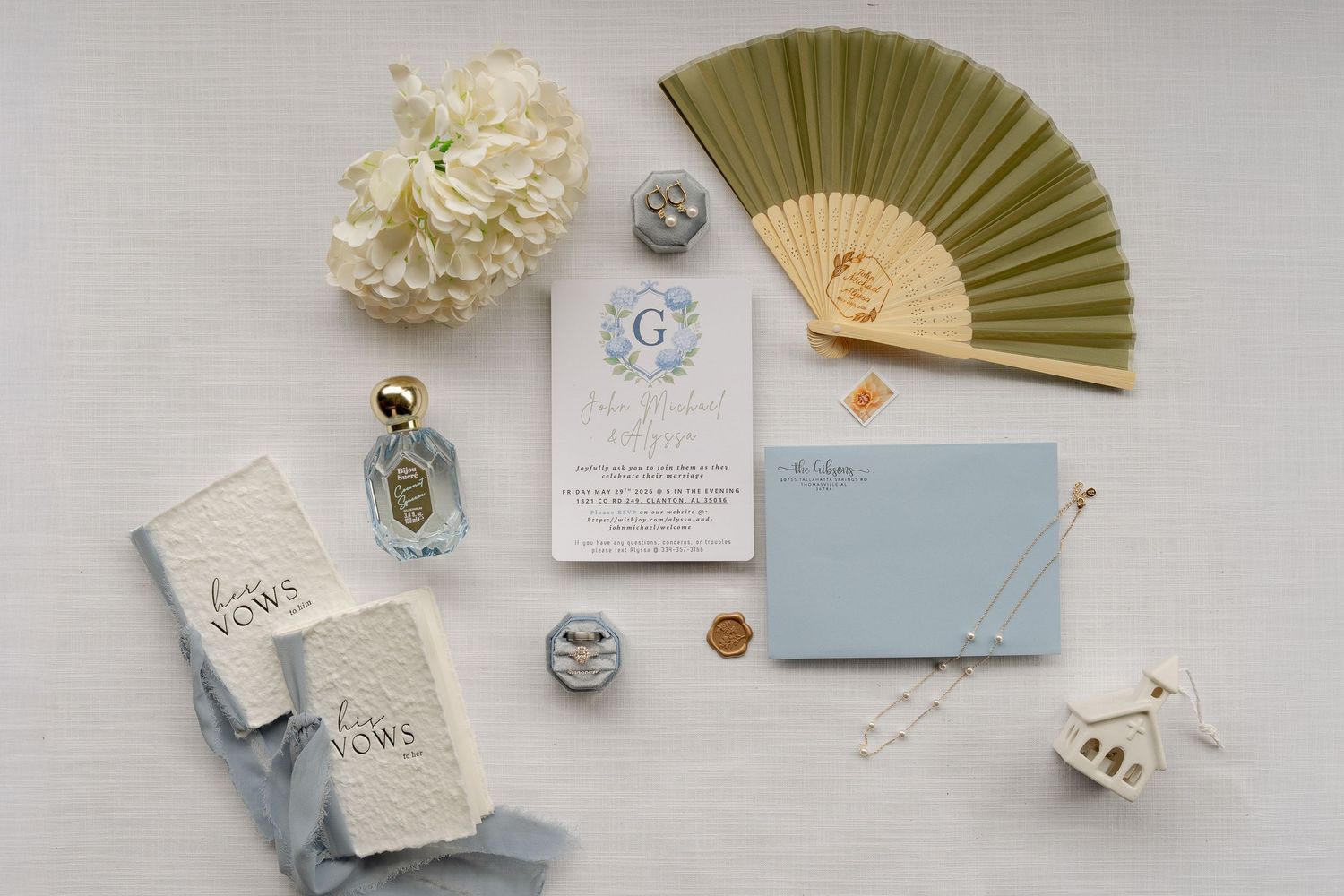 Elegant wedding flat lay with blue invitation suite, gold wax seal, vow books, perfume bottle, and olive green paper fan.