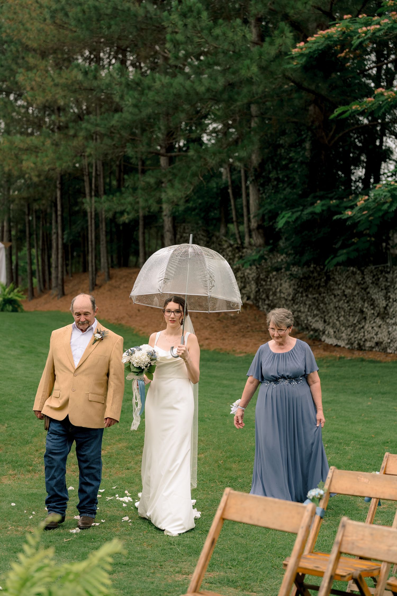 Bride walks down outdoor aisle with parents holding clear umbrella in white gown, bouquet, green trees backdrop.