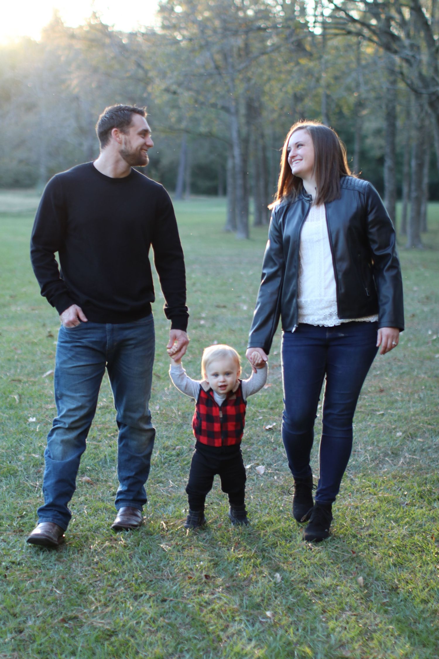 Family walking together outdoors with toddler in red plaid vest holding hands between parents.