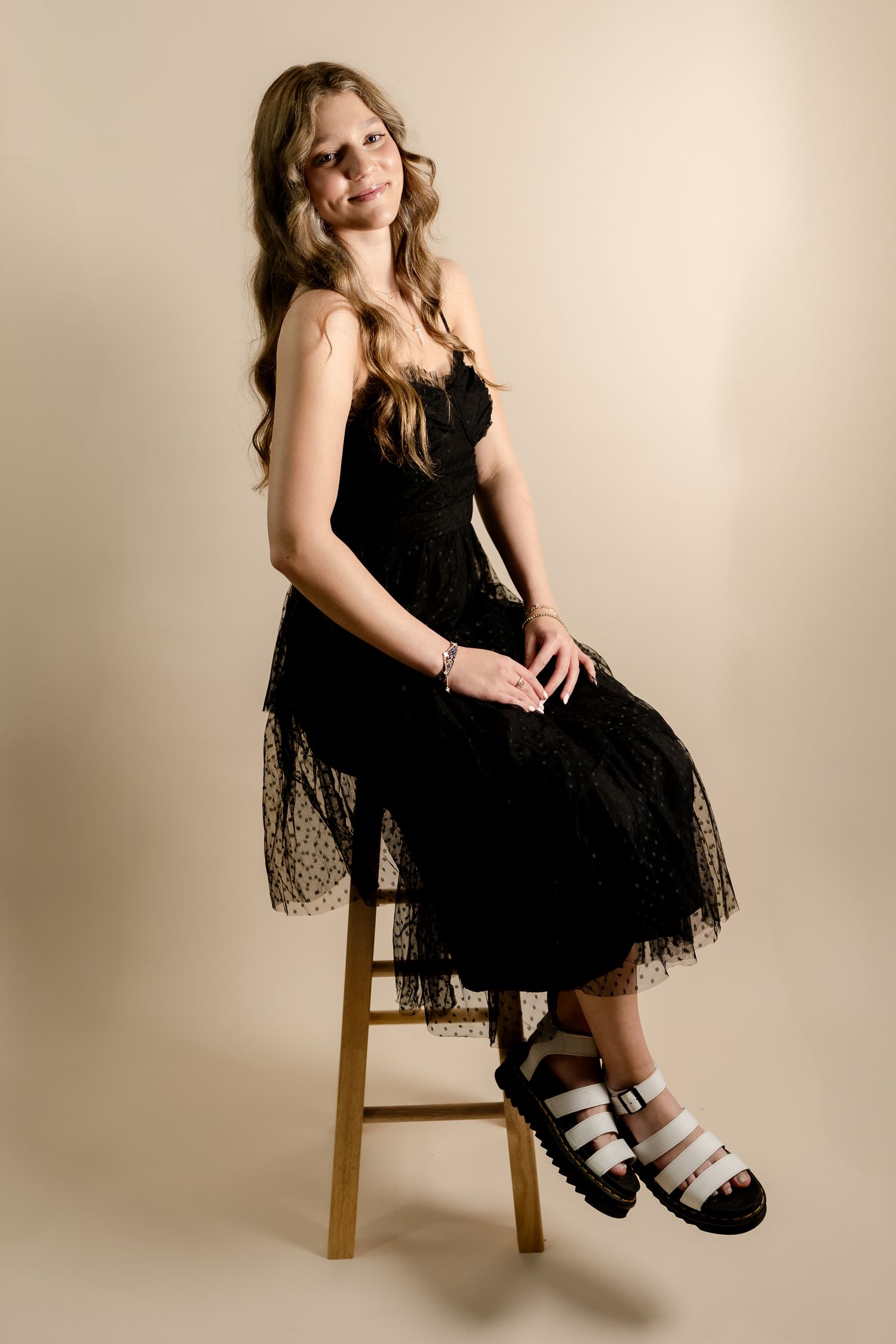 Woman in black dress sits elegantly on wooden stool against beige backdrop.