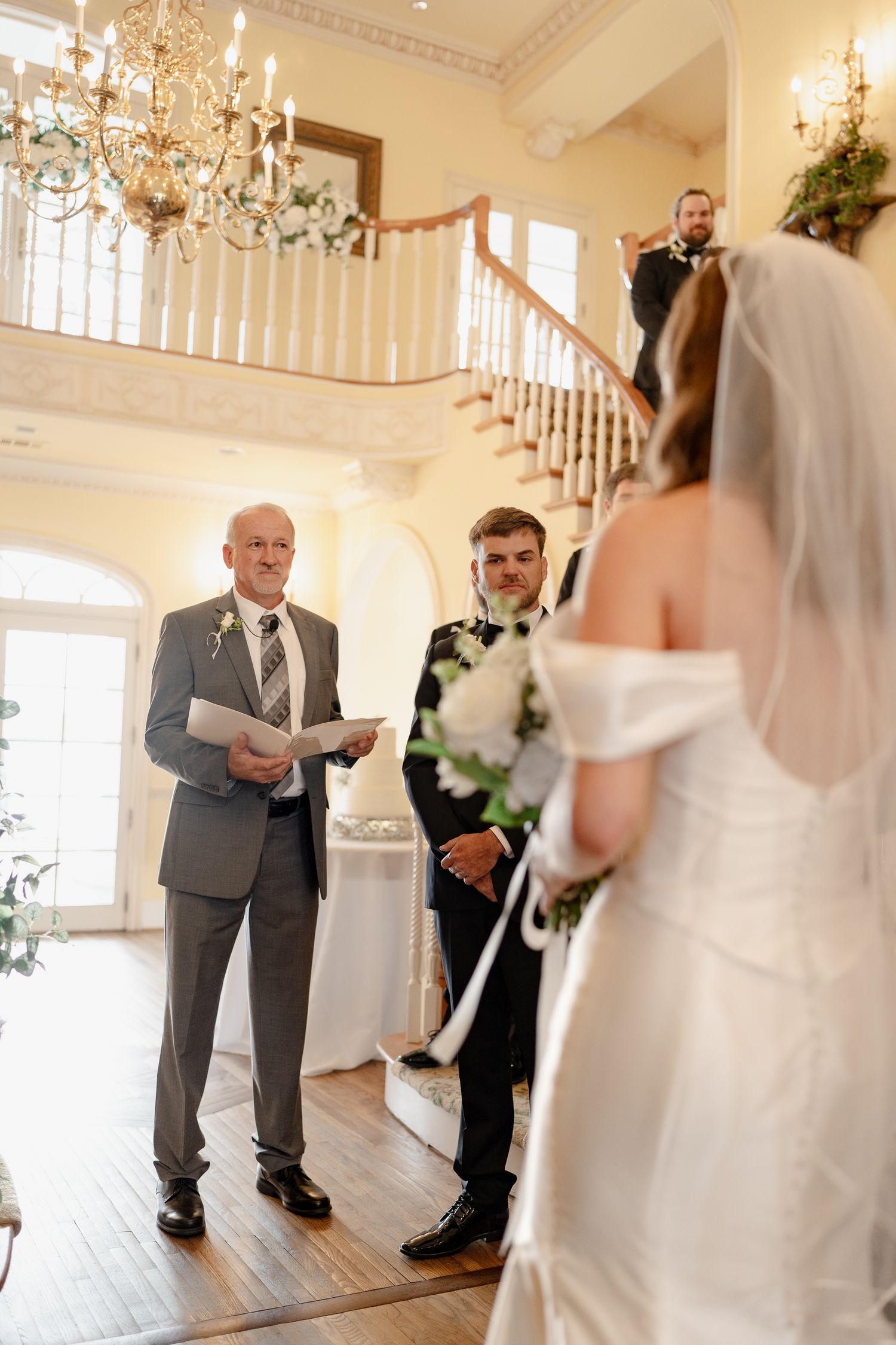 Wedding ceremony in progress in an elegant room with chandelier and white staircase.