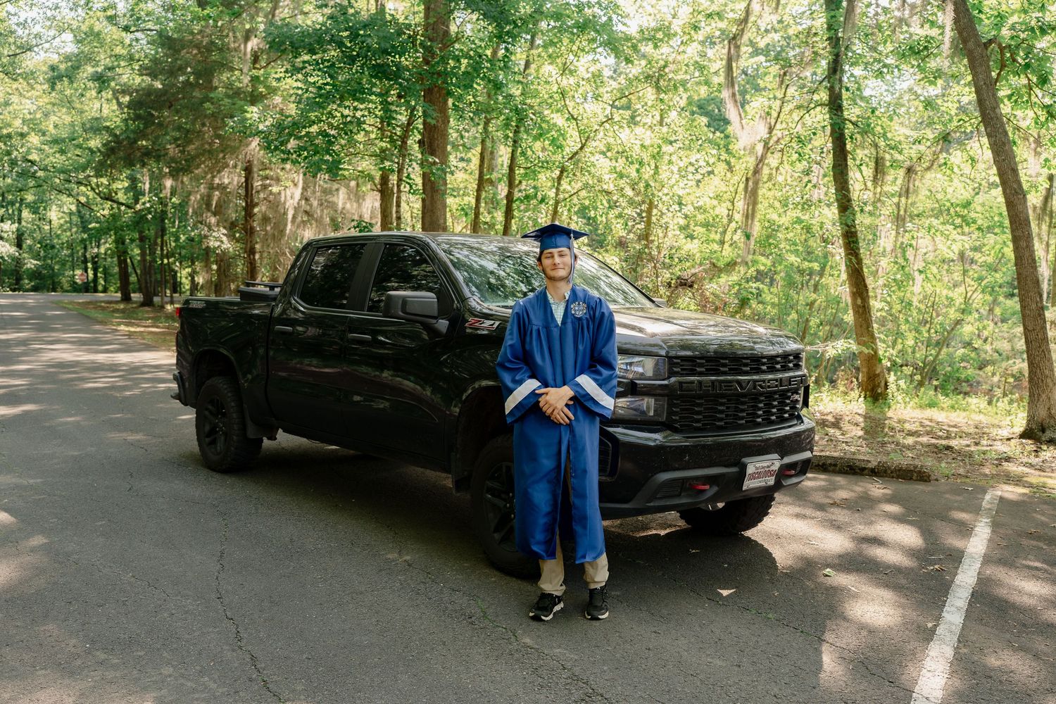 Graduate in academic regalia poses with black GMC pickup truck in wooded setting.