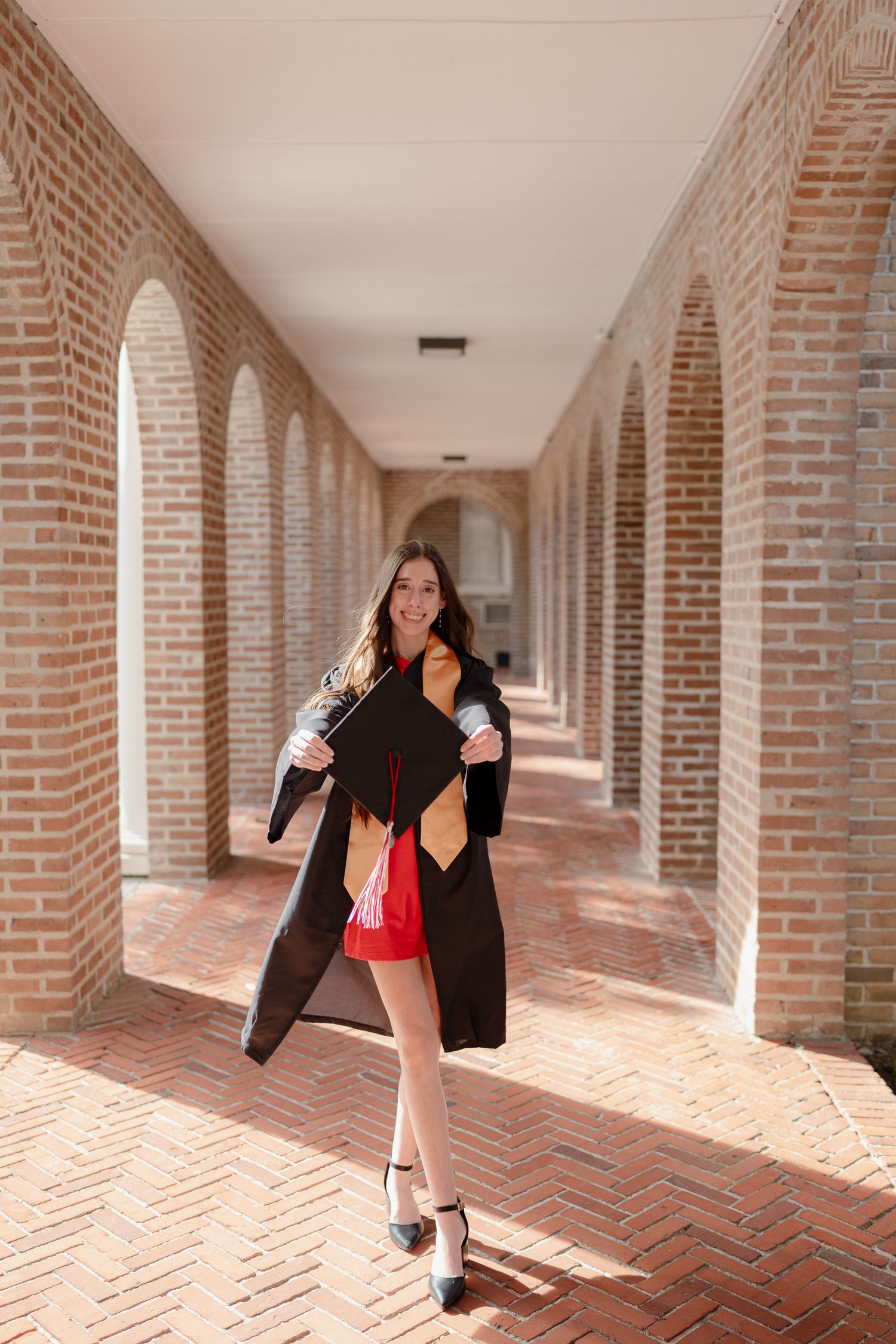 Woman in black dress and boots walks through a sunlit brick colonnade with arched columns and herringbone pattern floor.
