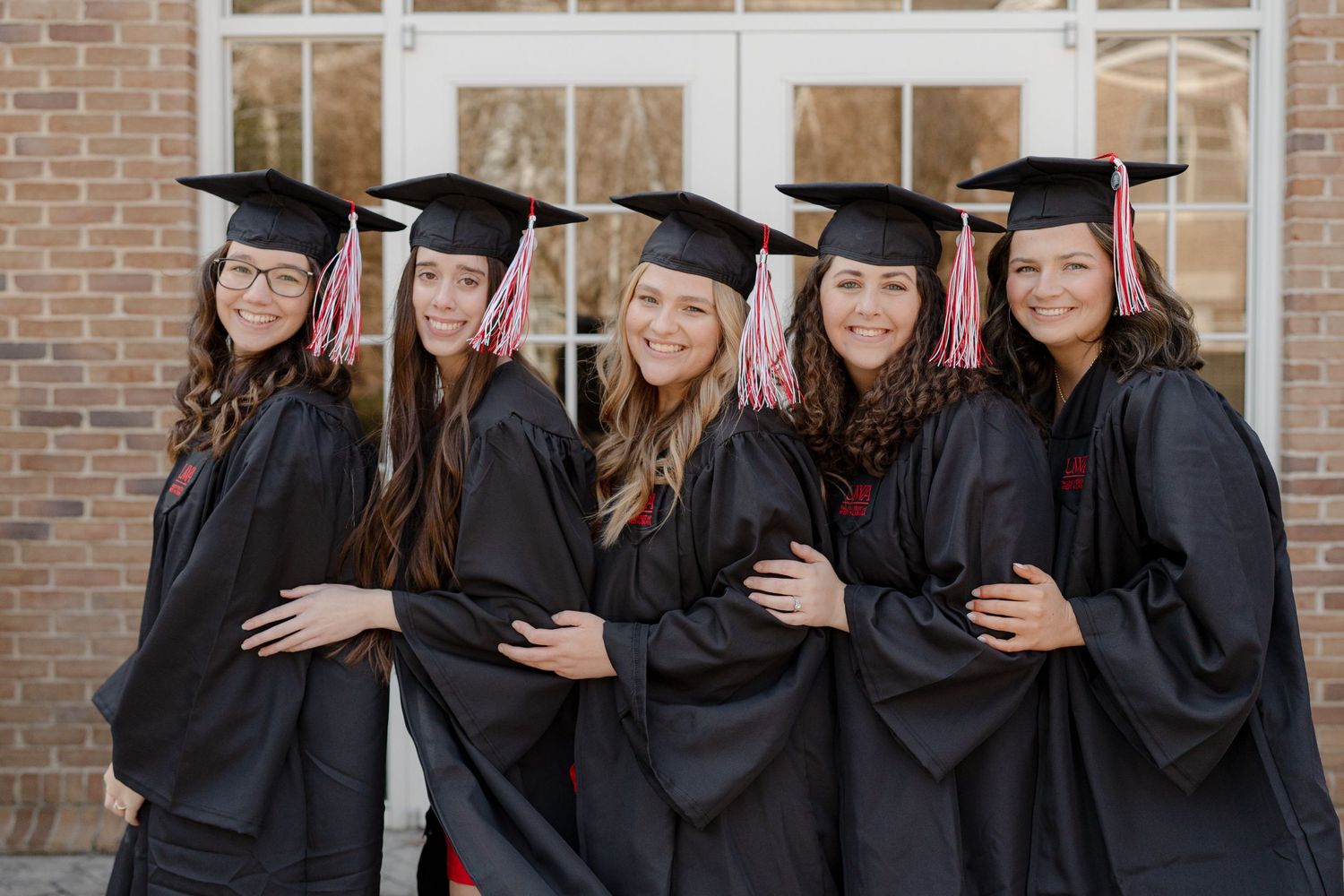 A group of graduates in black caps and gowns pose together against a brick wall during a graduation ceremony.