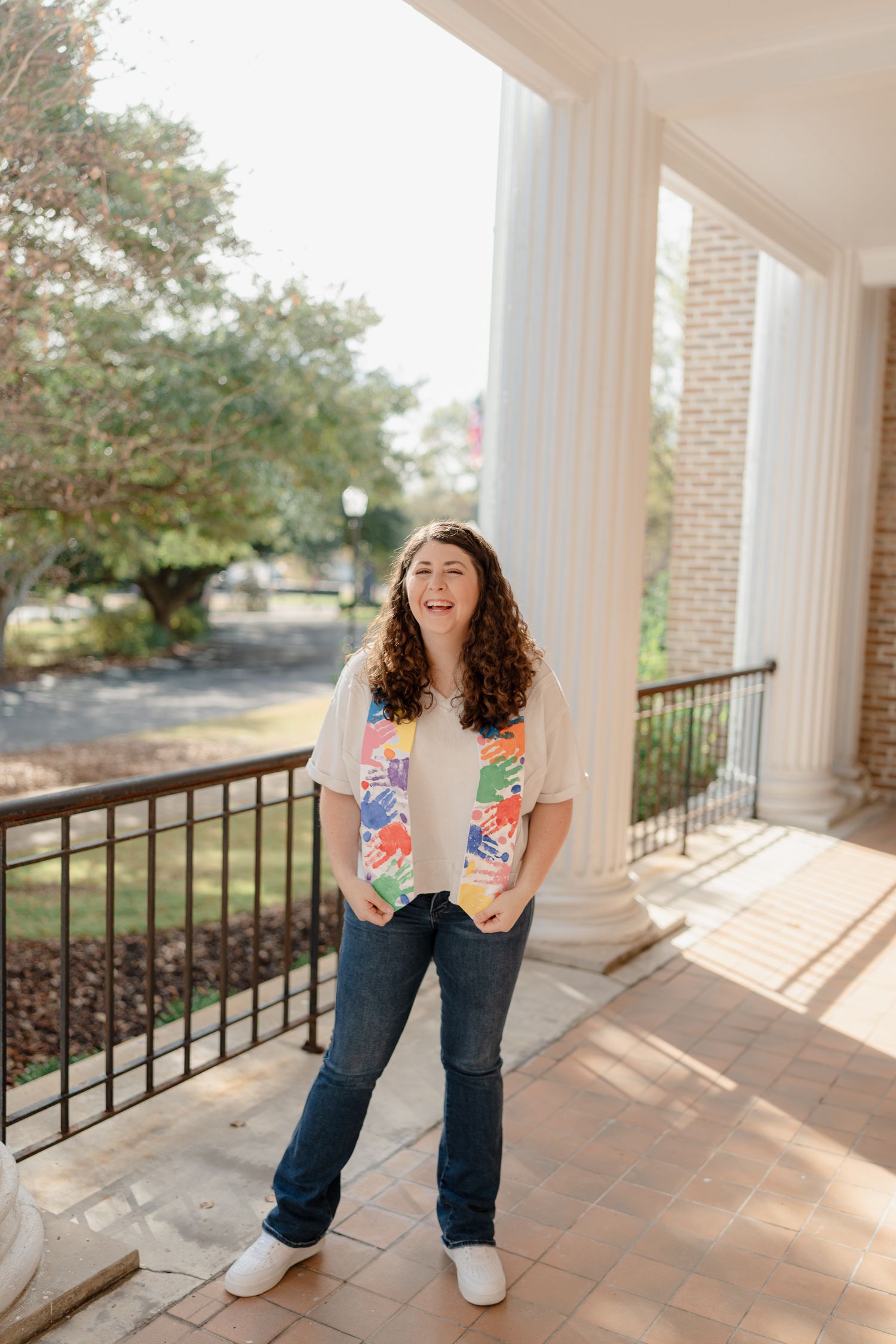 A person in jeans and a floral top stands laughing on a covered porch with white columns and brick flooring.