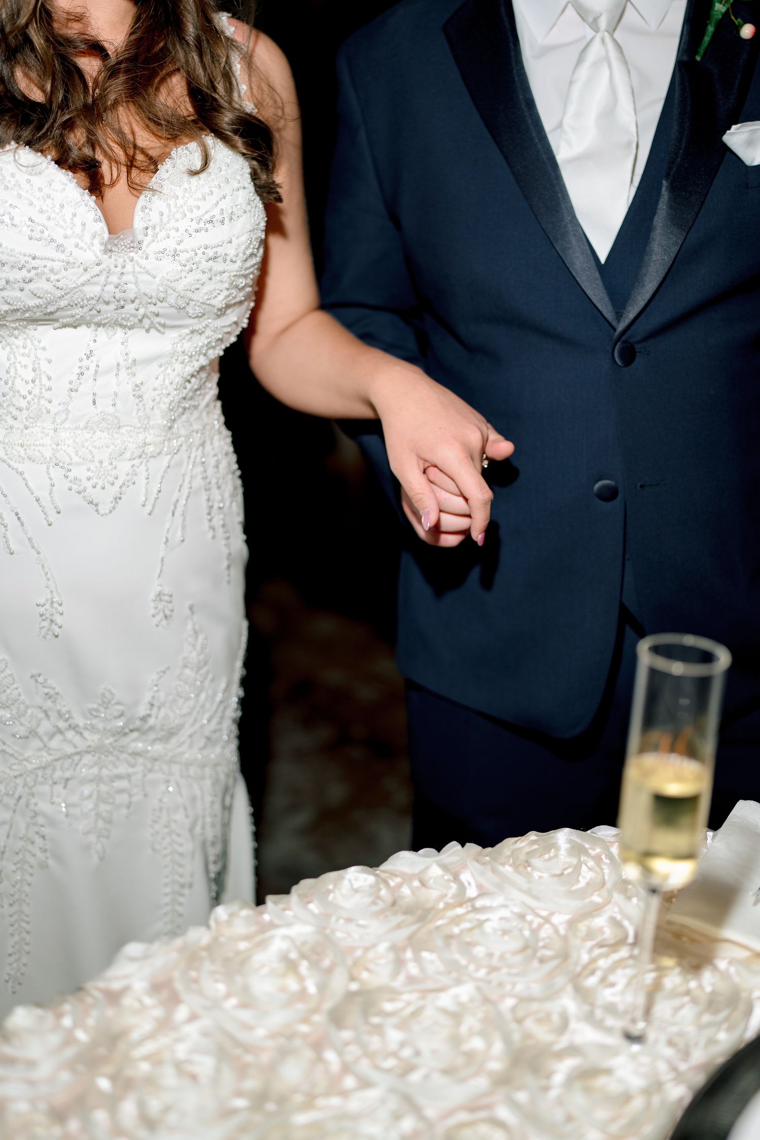 Bride and groom hold hands near white wedding cake, bride in beaded gown, groom in navy suit.