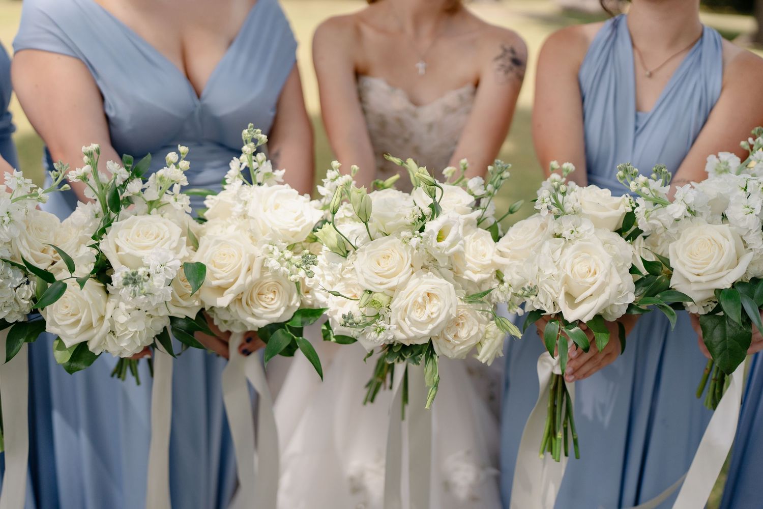 A row of bridesmaids in dusty blue dresses hold elegant white and cream floral bouquets at a wedding ceremony.